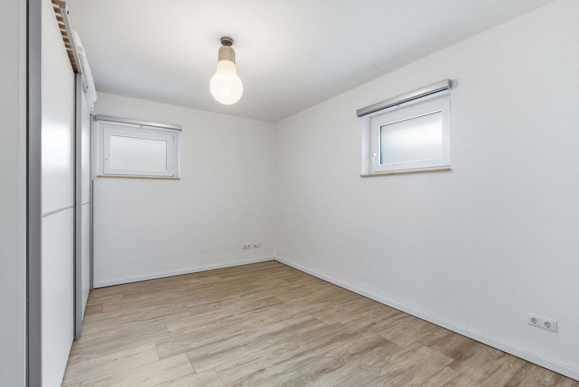Leeres Zimmer mit Holzfußboden, zwei kleinen Fenstern und weißer Decke mit Lampe.