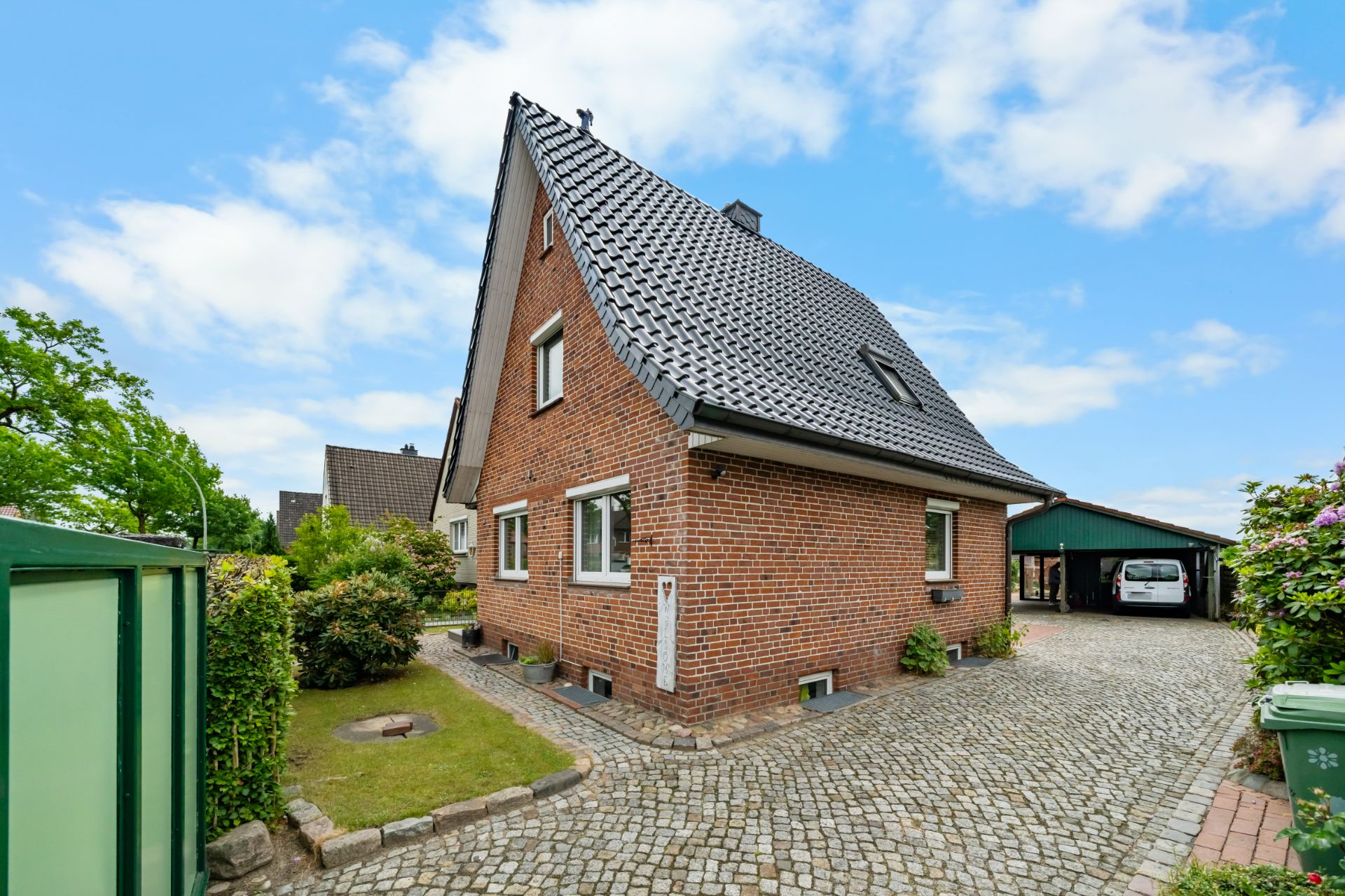 Backsteinwohnhaus mit steilem Dach, gepflasterte Einfahrt und Carport im Garten.