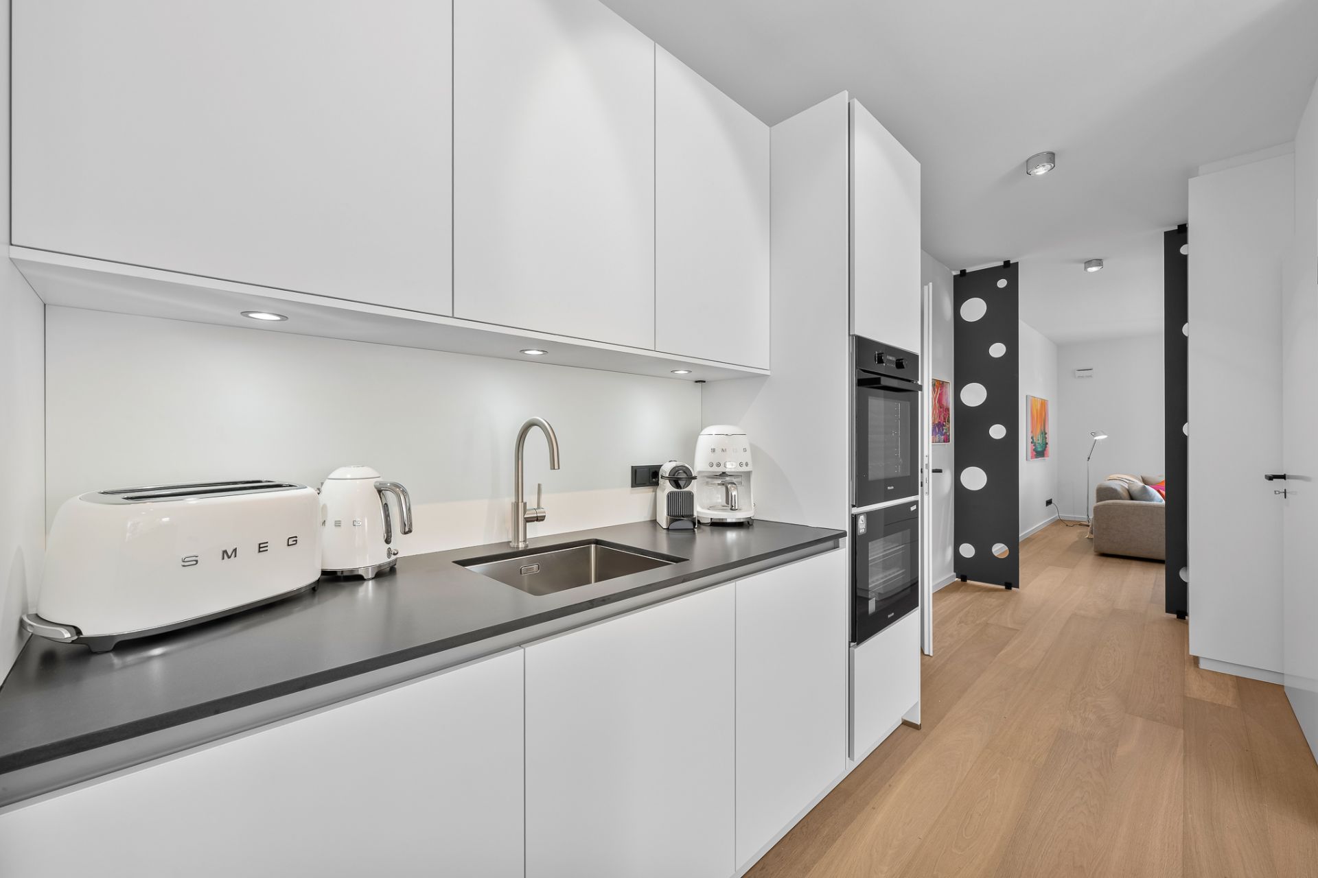 Moderne weiße Einbauküche mit Spüle, Toaster, Wasserkocher und Kaffeemaschine auf dunkler Arbeitsfläche.