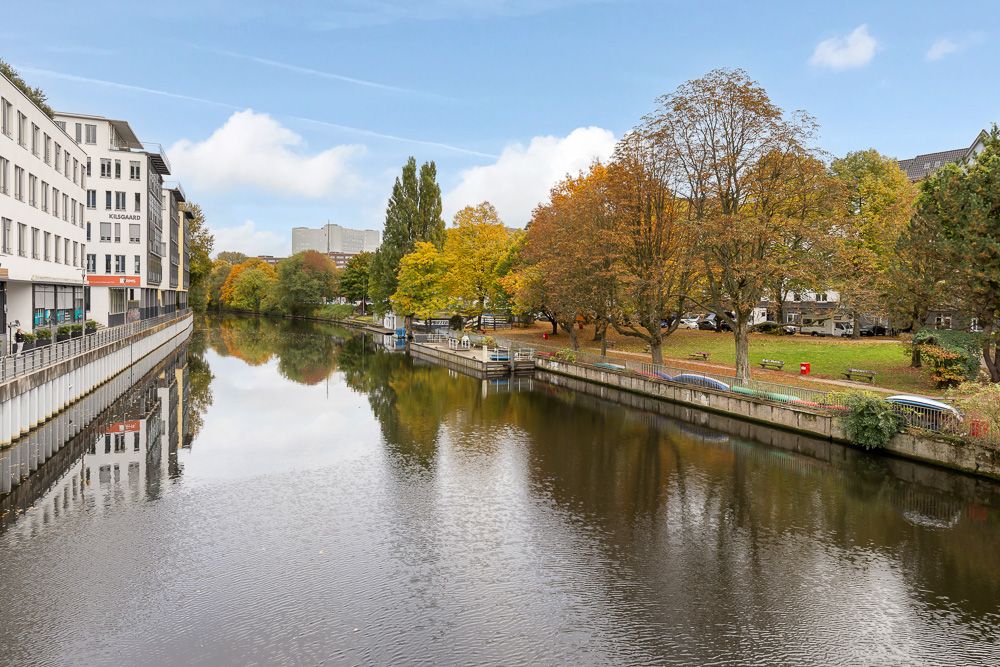 Fluss mit Uferbäumen und mehrstöckigem Wohngebäude links im Bild.