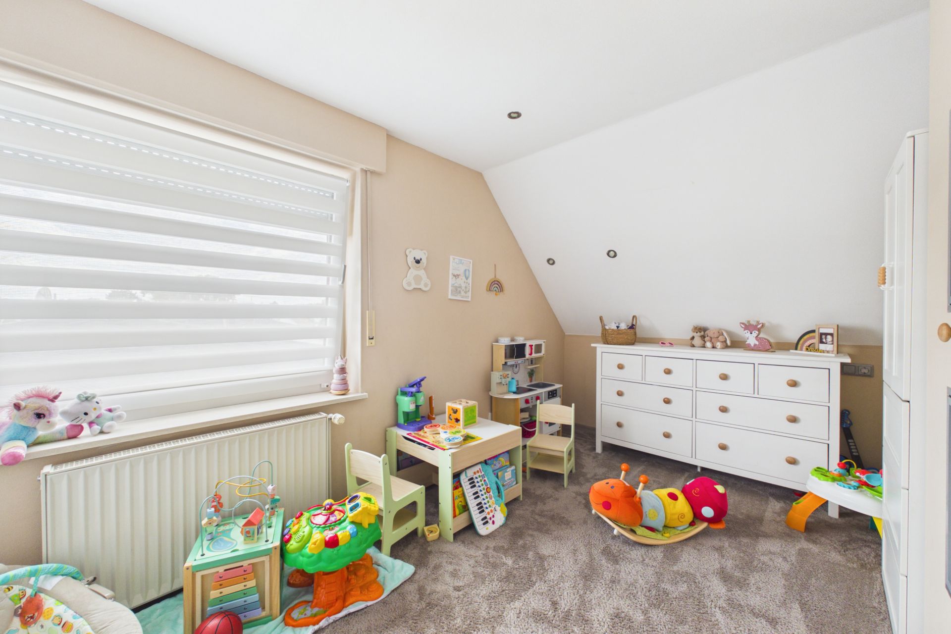 Helles Kinderzimmer mit Spielzeug, kleinem Tisch, Stühlen und weißer Kommode.