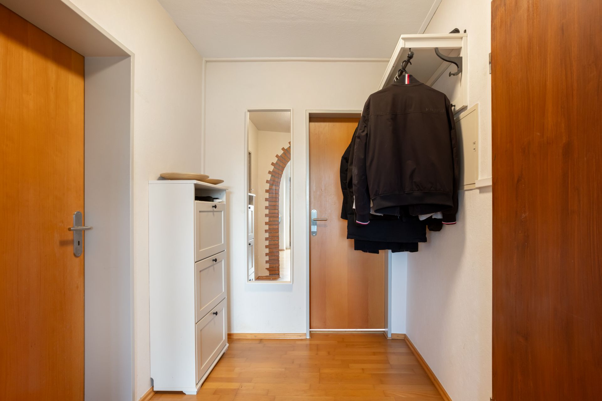 Flur mit Holztür, Spiegel, Schuhschrank und an der Wand hängenden Jacken.