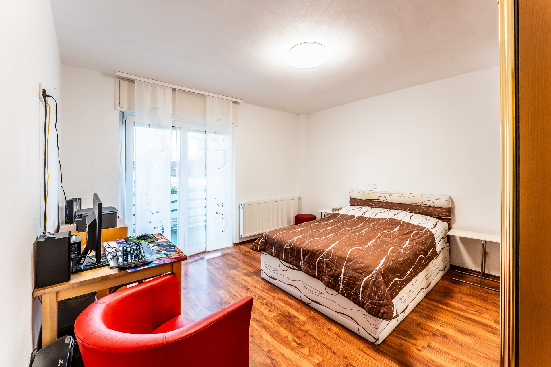 Schlafzimmer mit Doppelbett, Schreibtisch mit Computer und rotem Stuhl, Holzboden, Fenster mit Vorhang.
