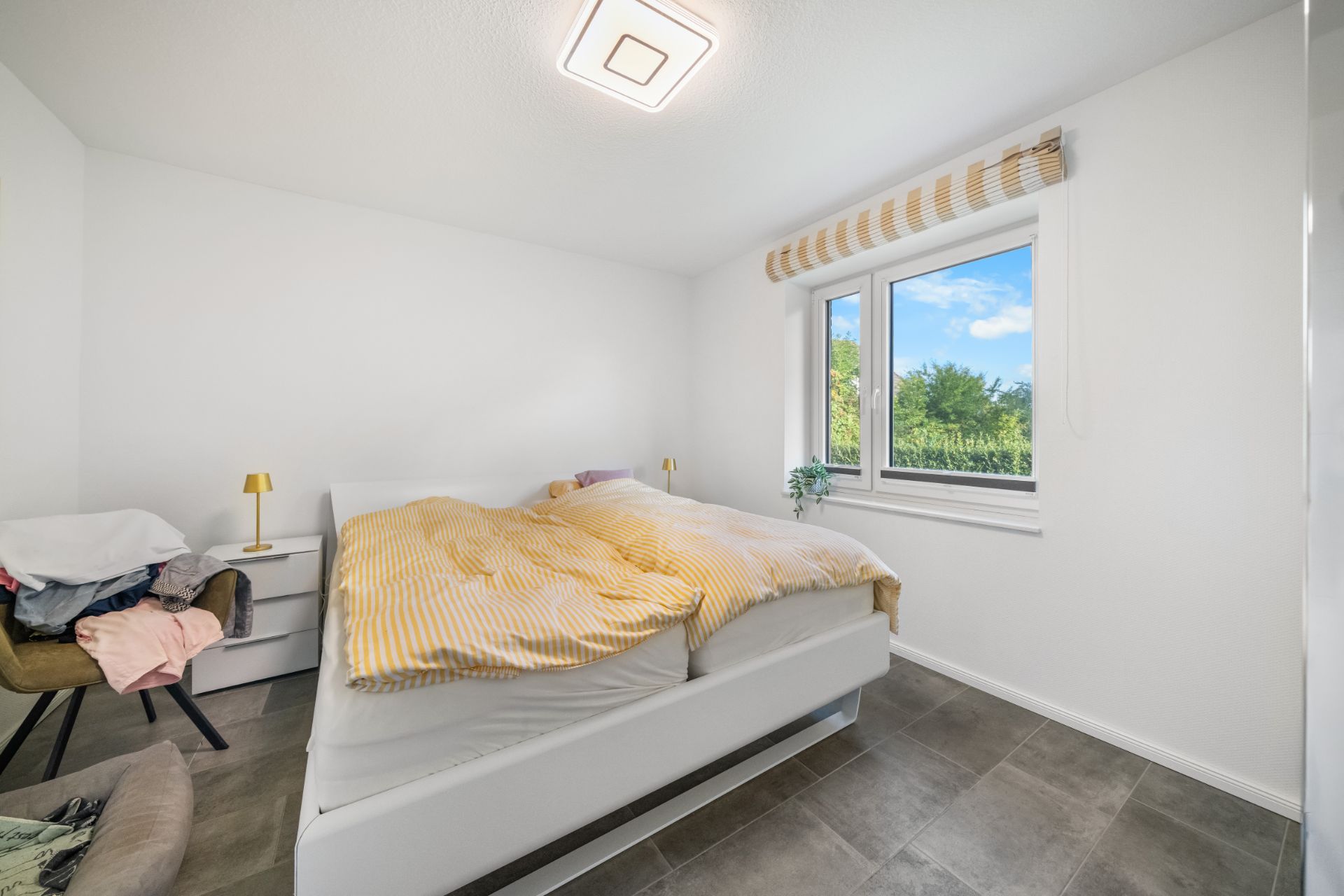 Helles Schlafzimmer mit Doppelbett, Fenster, Nachttischen und einem Stuhl mit Kleidung.