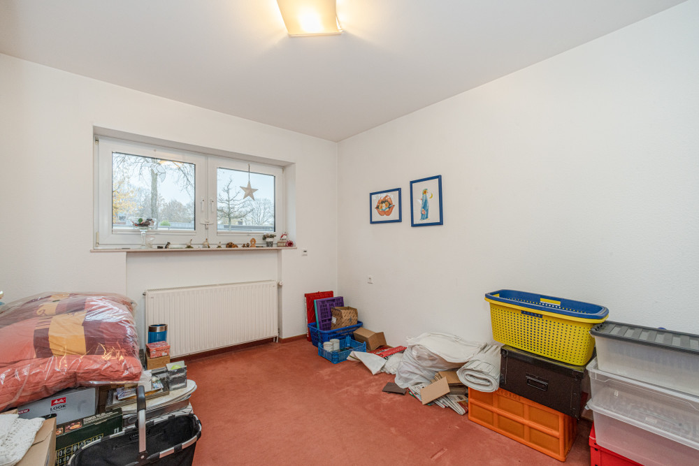 Kleines Zimmer mit Teppichboden, Fenster, einigen Kisten und gestapelten Gegenständen an der Wand.