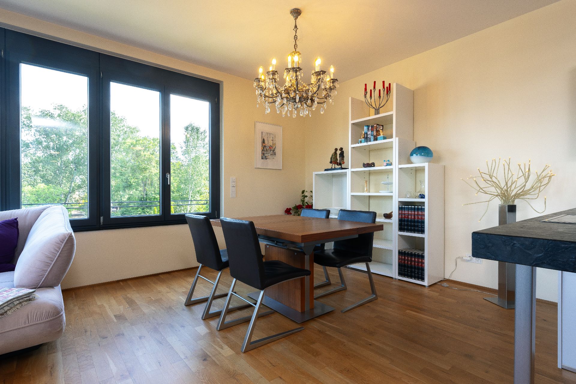 Essbereich mit Holztisch, vier schwarzen Stühlen, Kronleuchter und Regal vor großen Fenstern.