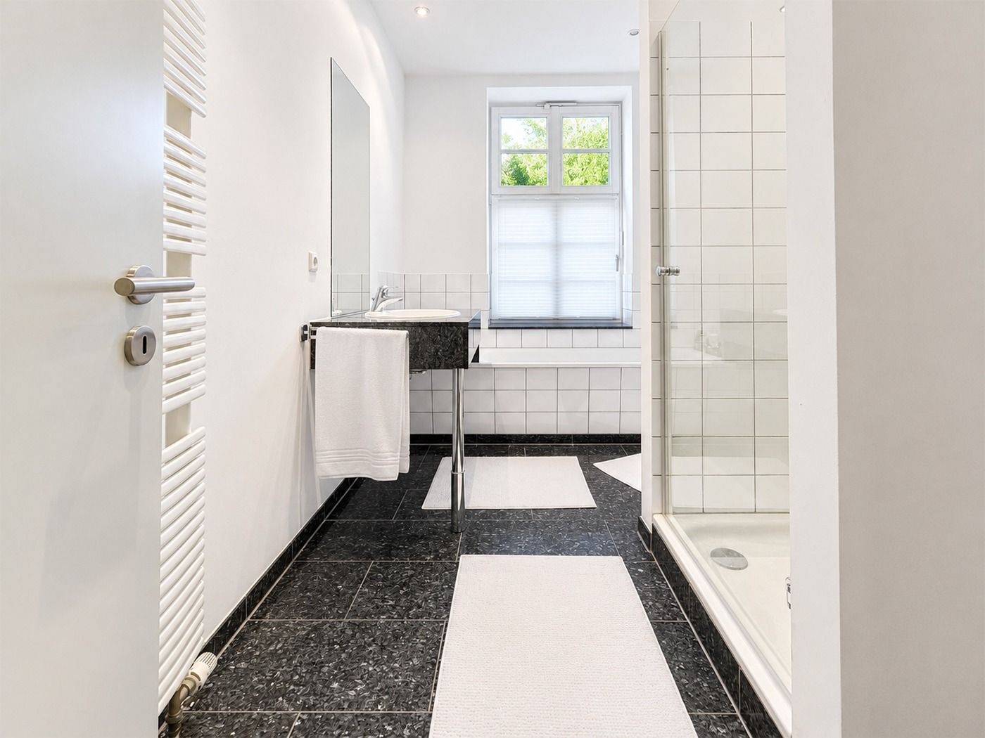 Helles Badezimmer mit Dusche, Waschbecken, Spiegel und Fenster, schwarze Fliesen und weiße Teppiche.