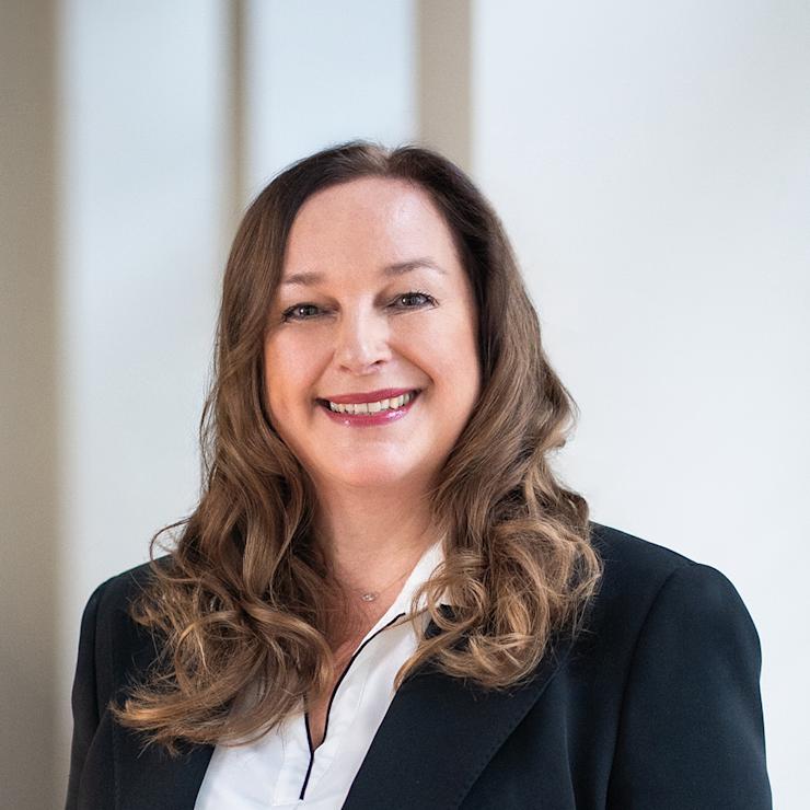 Frau mit langen braunen Haaren im Anzug lächelt in die Kamera.