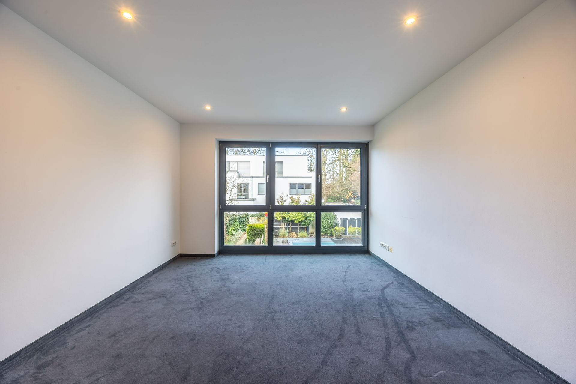 Leerer Raum mit grauem Teppichboden, weißen Wänden und großem Fenster mit Blick auf Garten und Nachbarhaus.