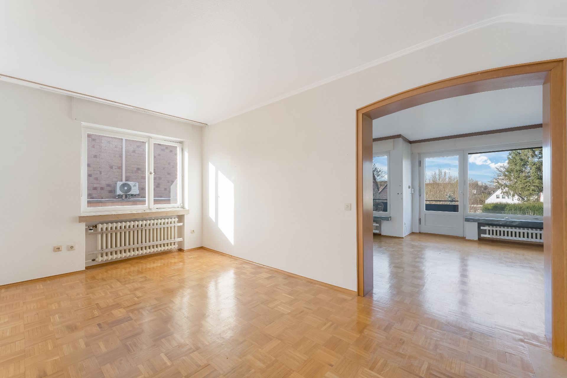 Helles, leeres Zimmer mit Parkettboden und großem Fenster, Durchgang zu weiterem Raum.