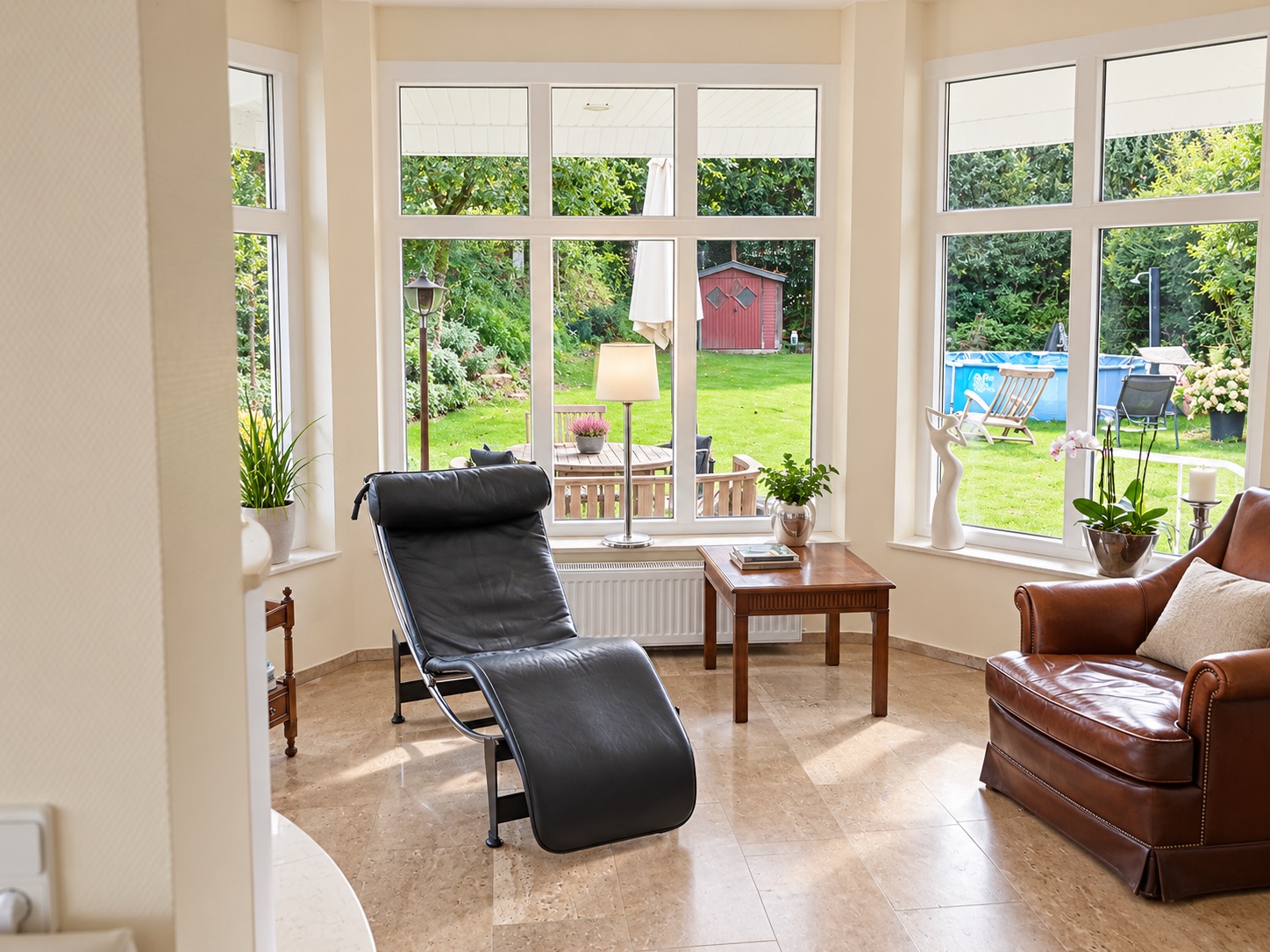 Heller Wohnbereich mit Liegesessel, Ledersessel, Tisch und großen Fenstern mit Blick in den Garten.