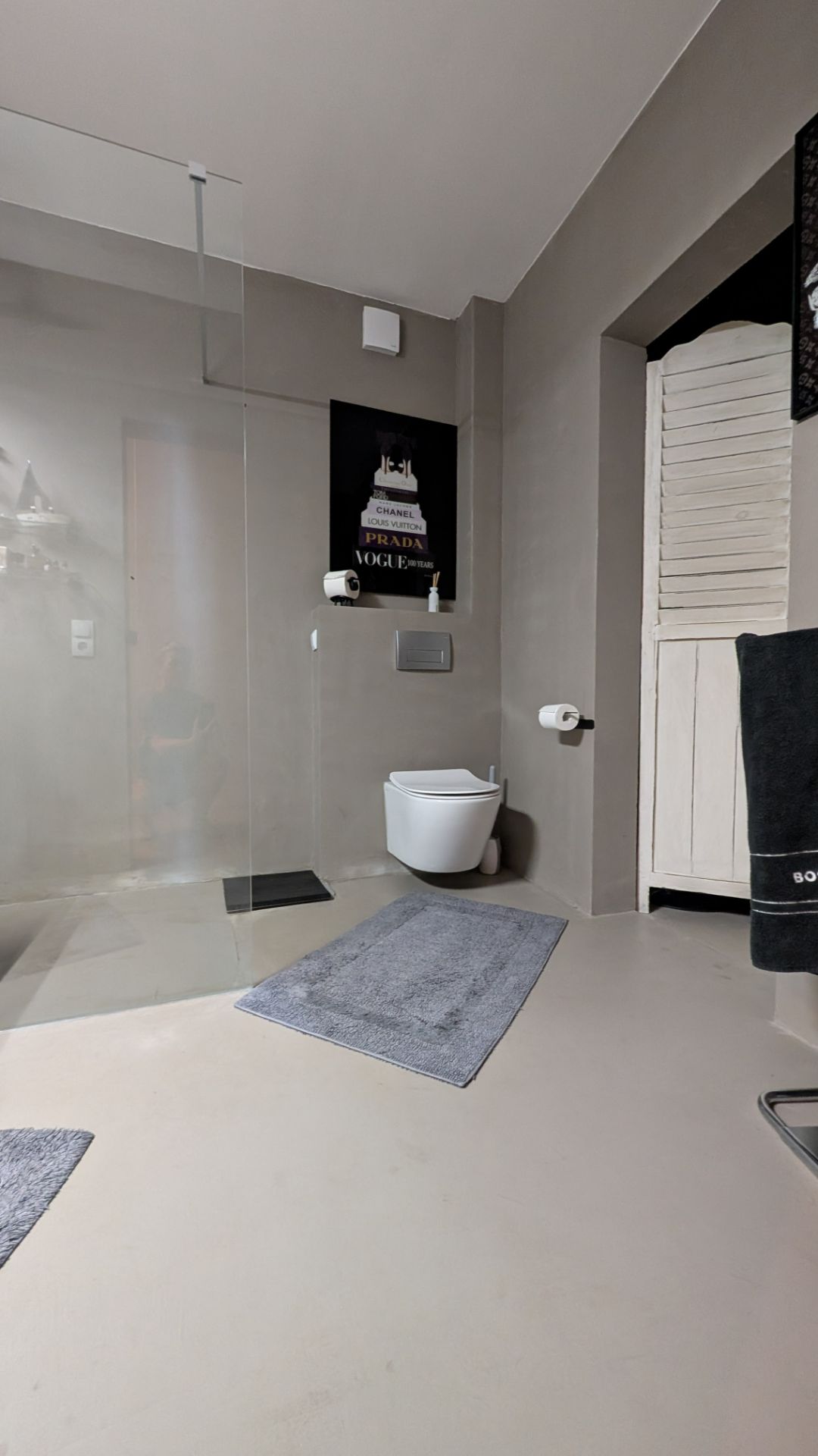 Modernes Badezimmer mit Wandtoilette, Glasdusche, grauen Teppichen und Handtüchern.
