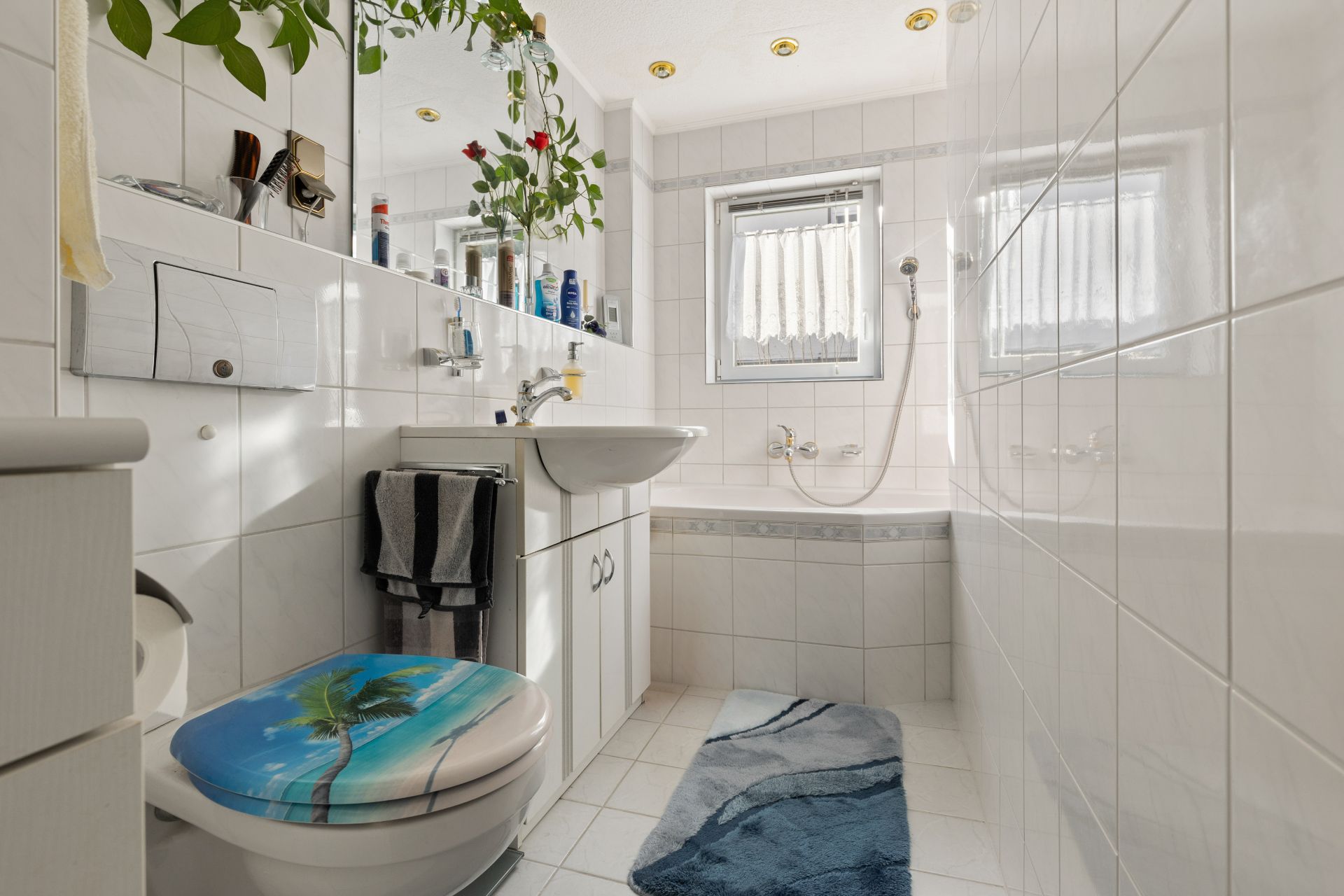 Helles Badezimmer mit Badewanne, Spiegel, Waschbecken und WC mit bemaltem Toilettendeckel.