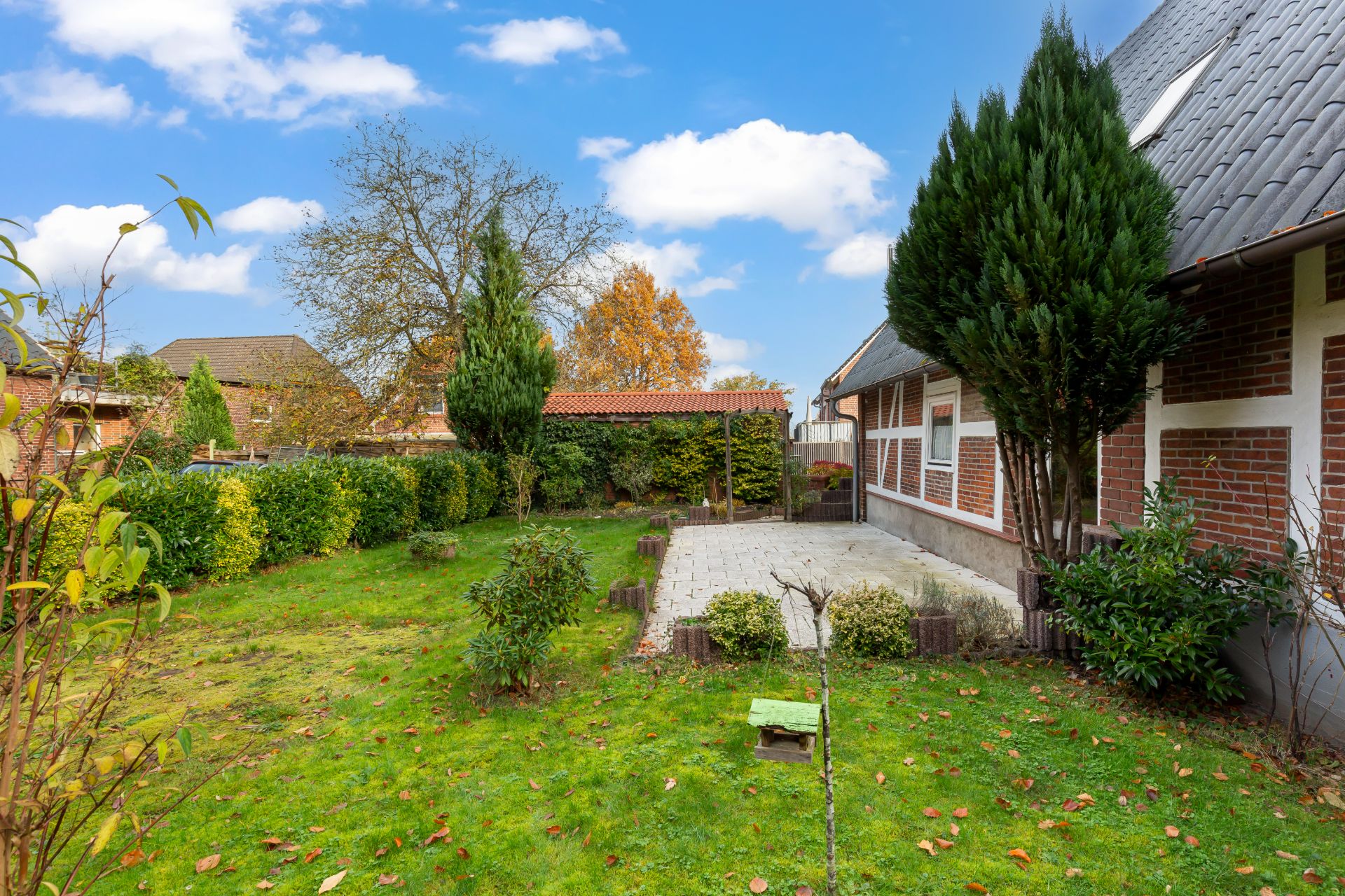 Grüner Garten mit Terrasse neben einem Fachwerkhaus, umgeben von Büschen und Bäumen.