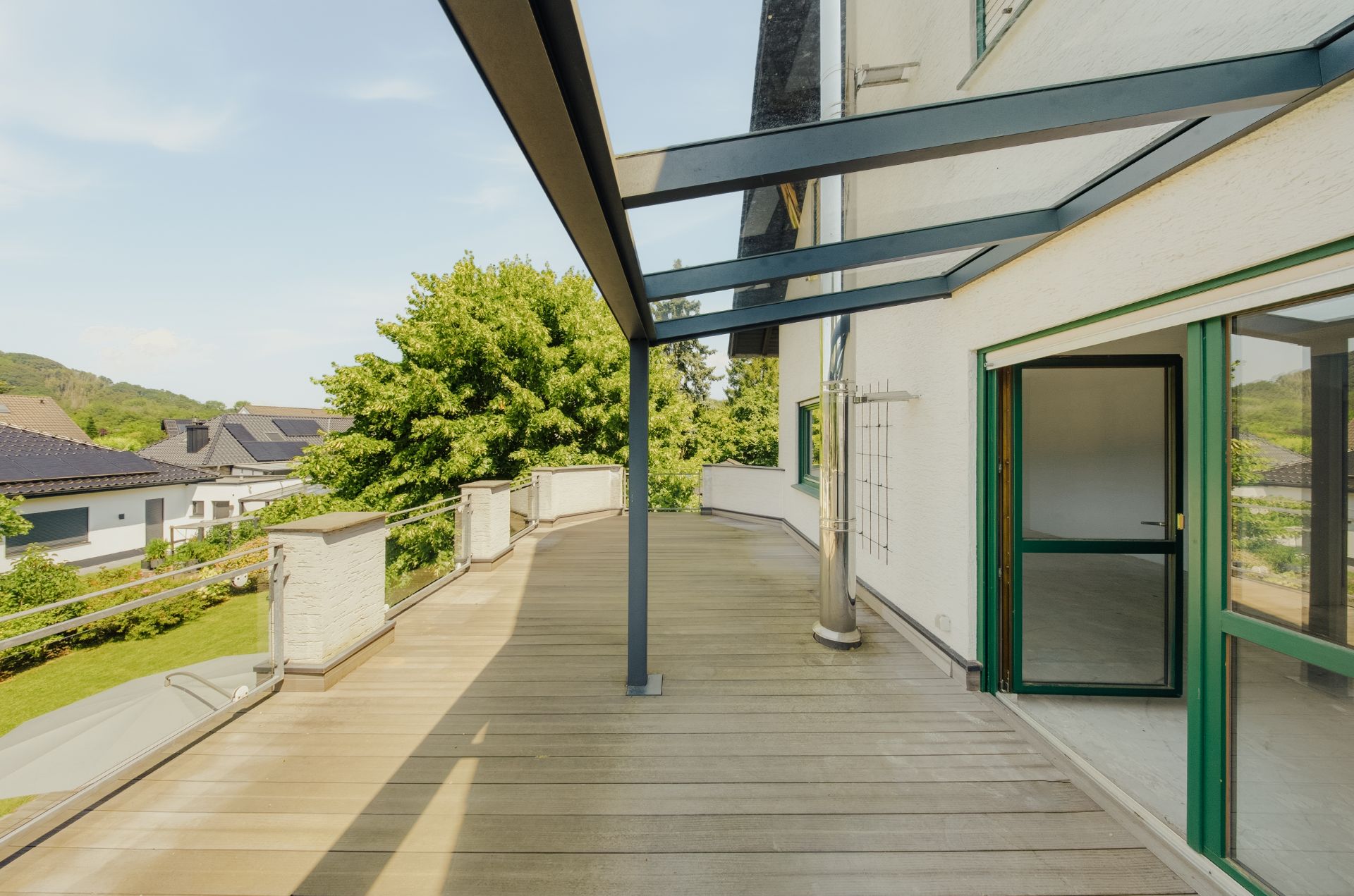 Große Terrasse mit Holzfußboden und Glasüberdachung, Zugang durch eine offene Tür, grüne Aussicht.