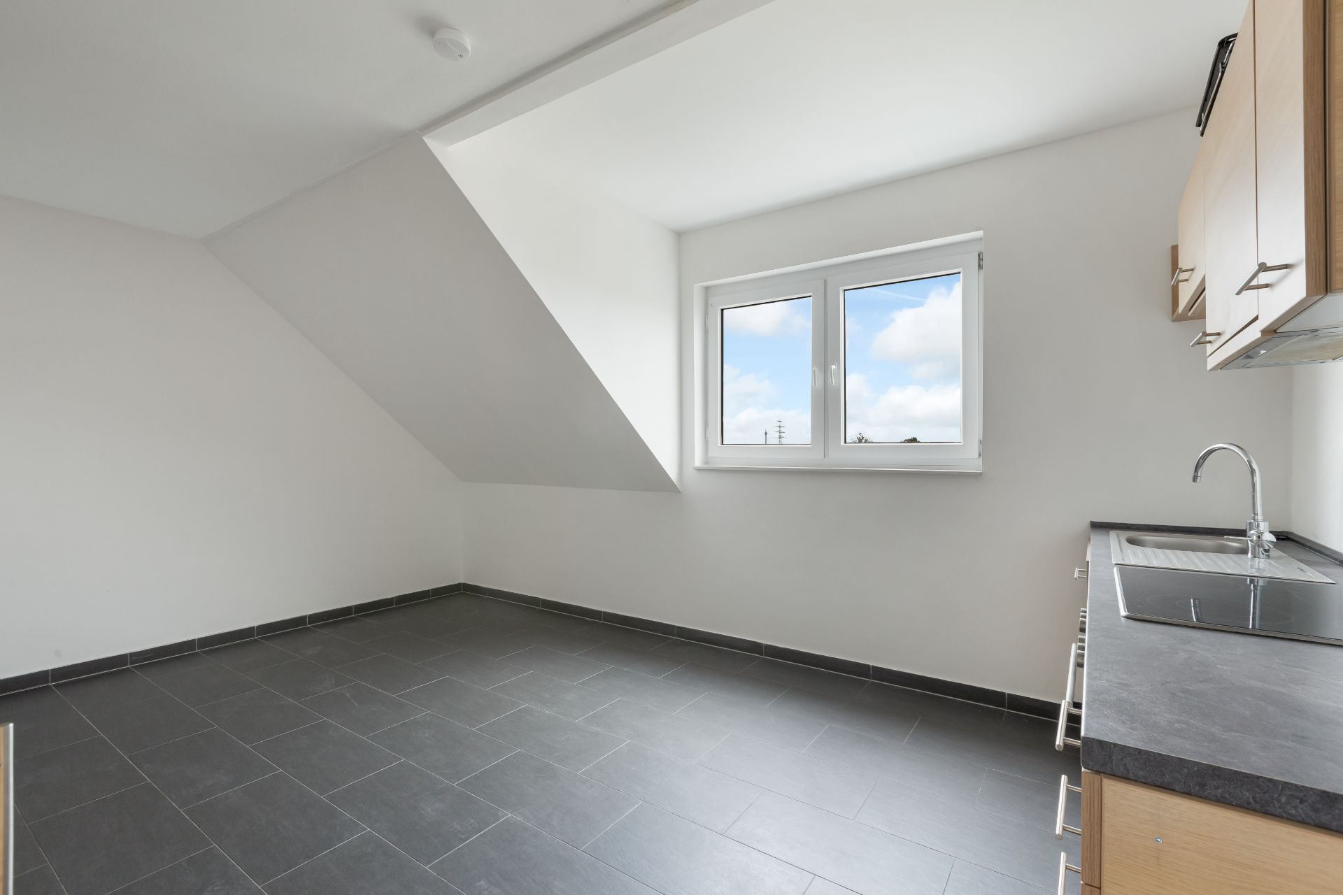 Leere Küche mit dunklem Fliesenboden, Fenster und Spüle auf der rechten Seite.