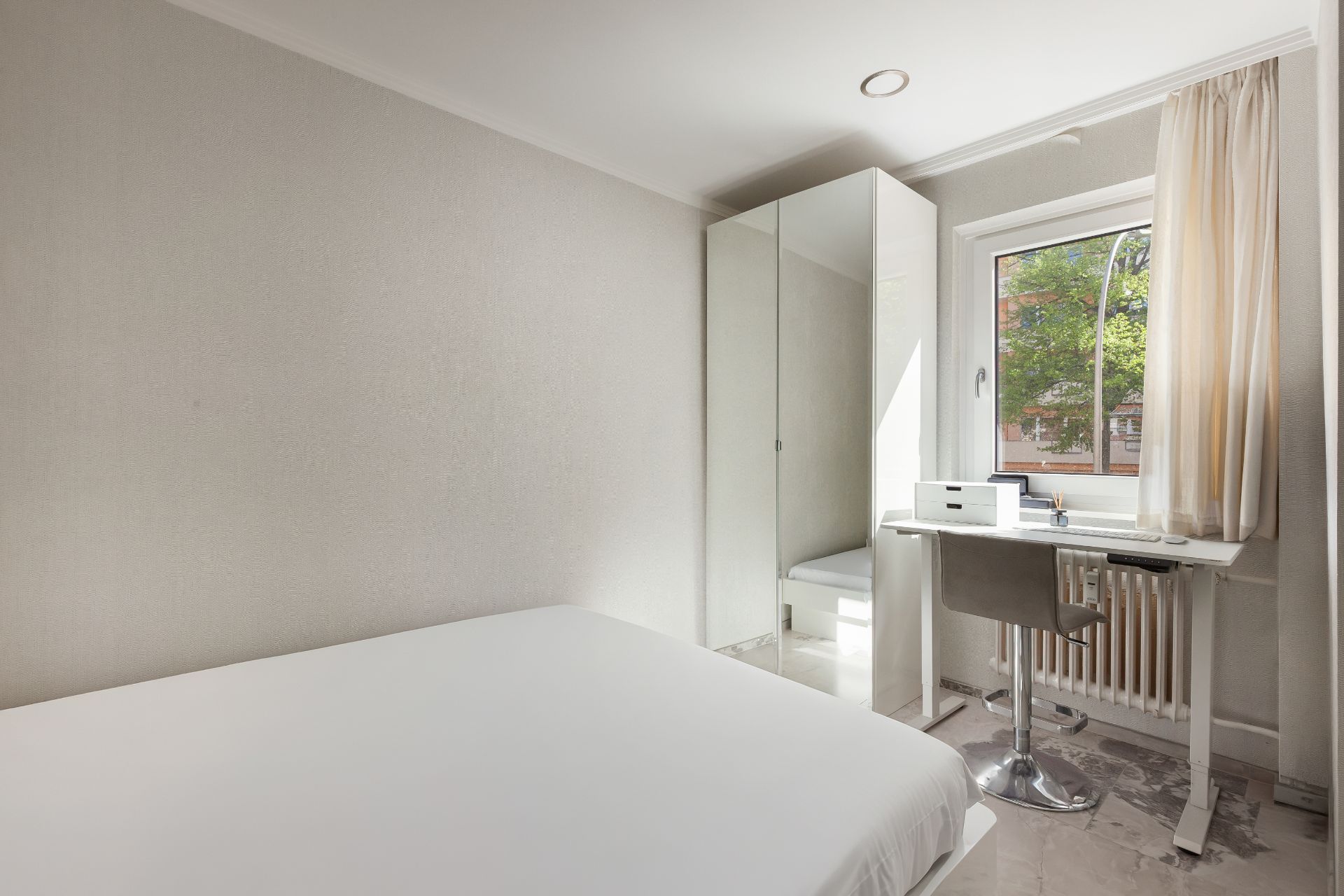 Helles Schlafzimmer mit Bett, Schreibtisch am Fenster, Stuhl und Kleiderschrank mit Spiegel.