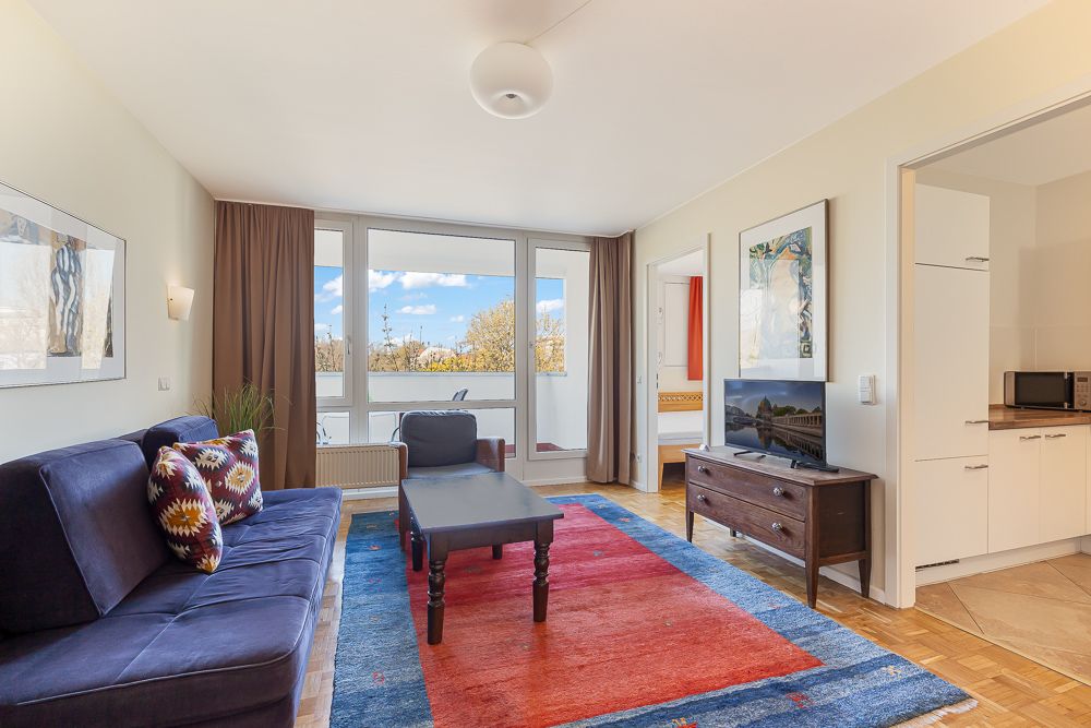Helles Wohnzimmer mit Sofa, Tisch, TV, buntem Teppich und Zugang zu einem Balkon.