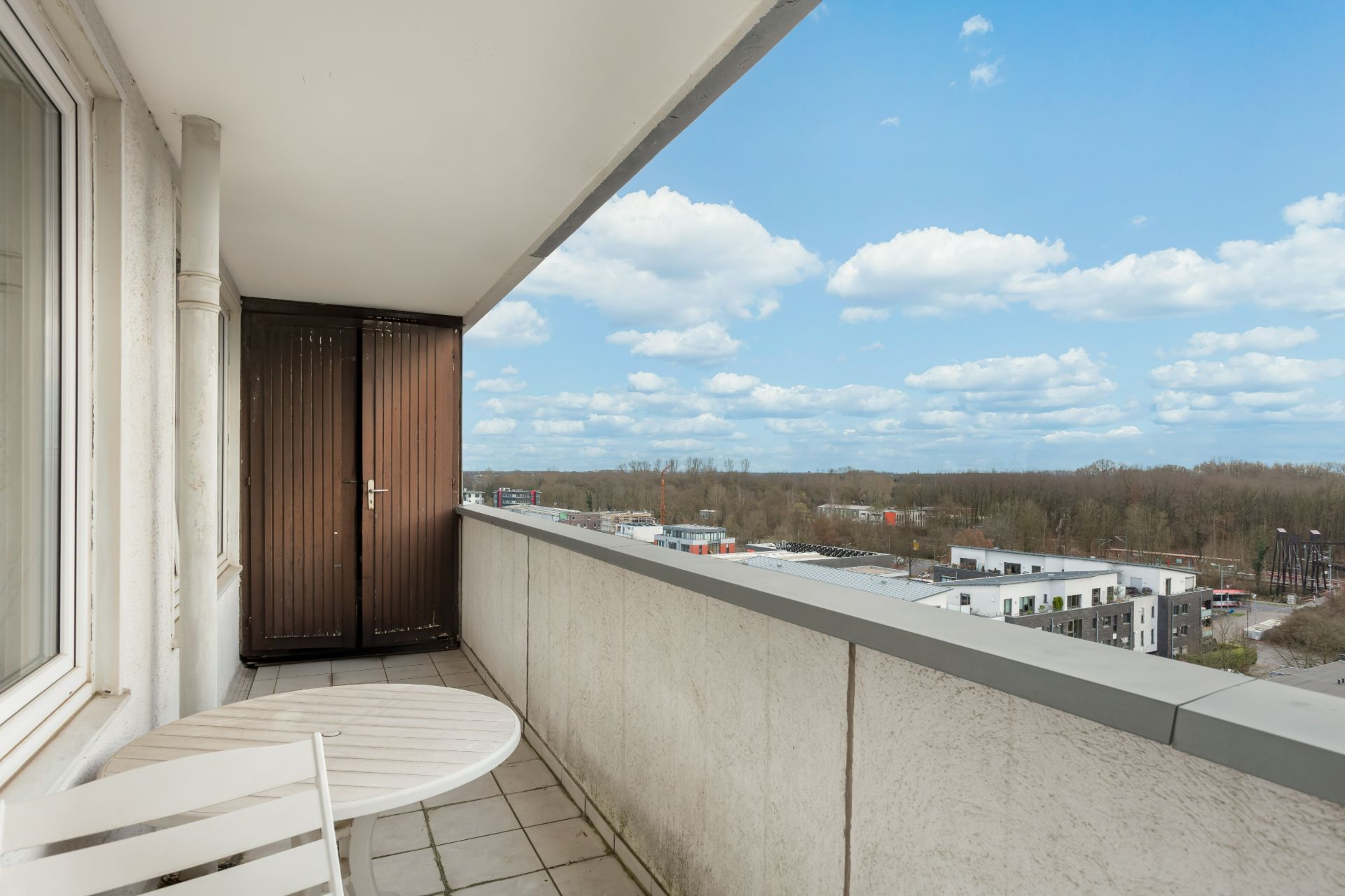 Balkon mit kleinem Tisch und Stuhl, Blick auf Gebäude und Bäume im Hintergrund.