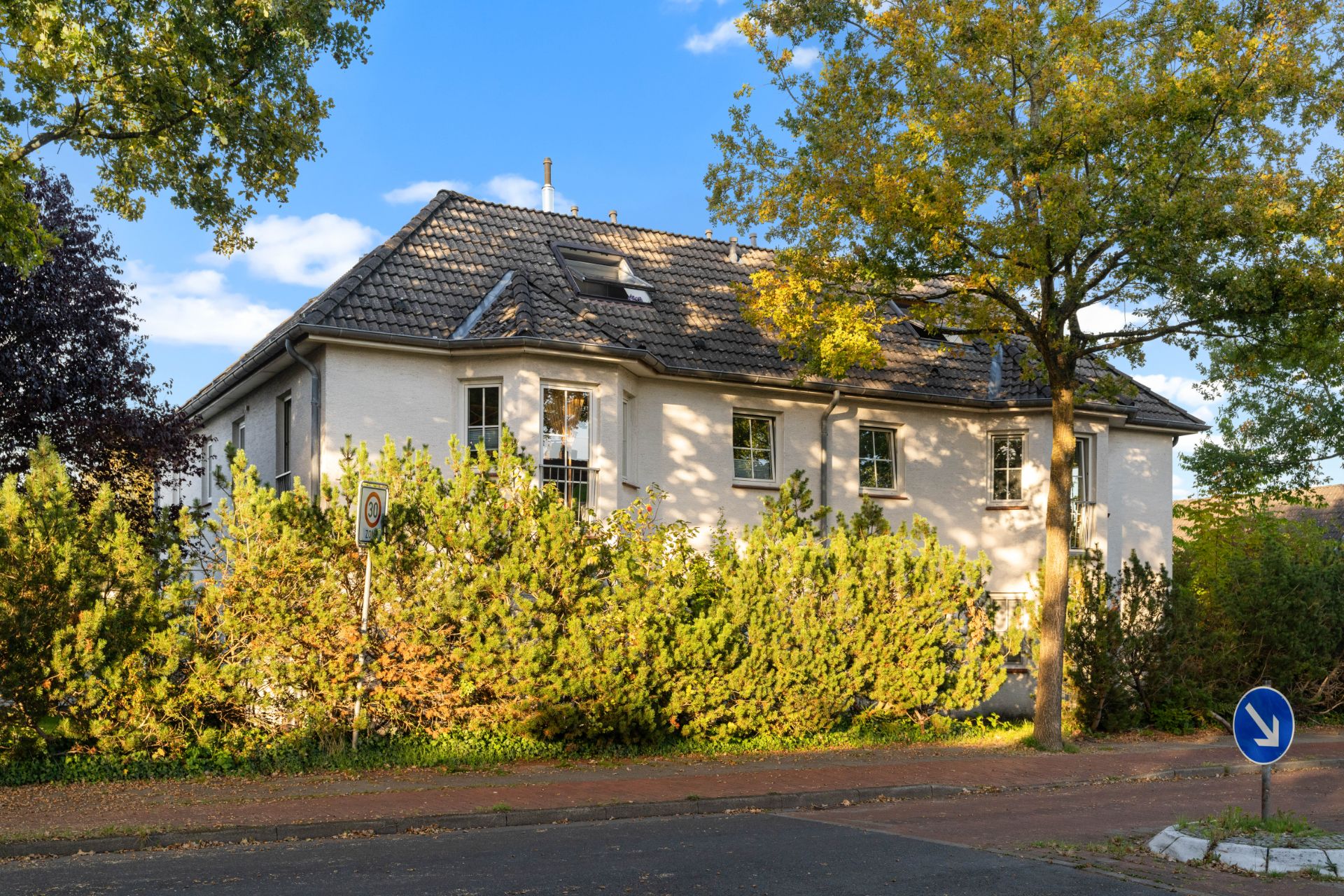 Zweistöckiges Wohnhaus mit Dachfenstern, umgeben von Bäumen und Sträuchern an einer ruhigen Straße.