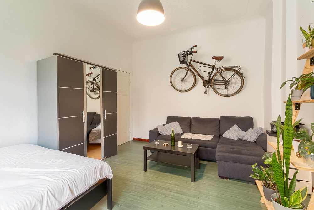 Wohnzimmer mit grauem Sofa, Couchtisch, Schrank, Bett und Fahrrad an der Wand.