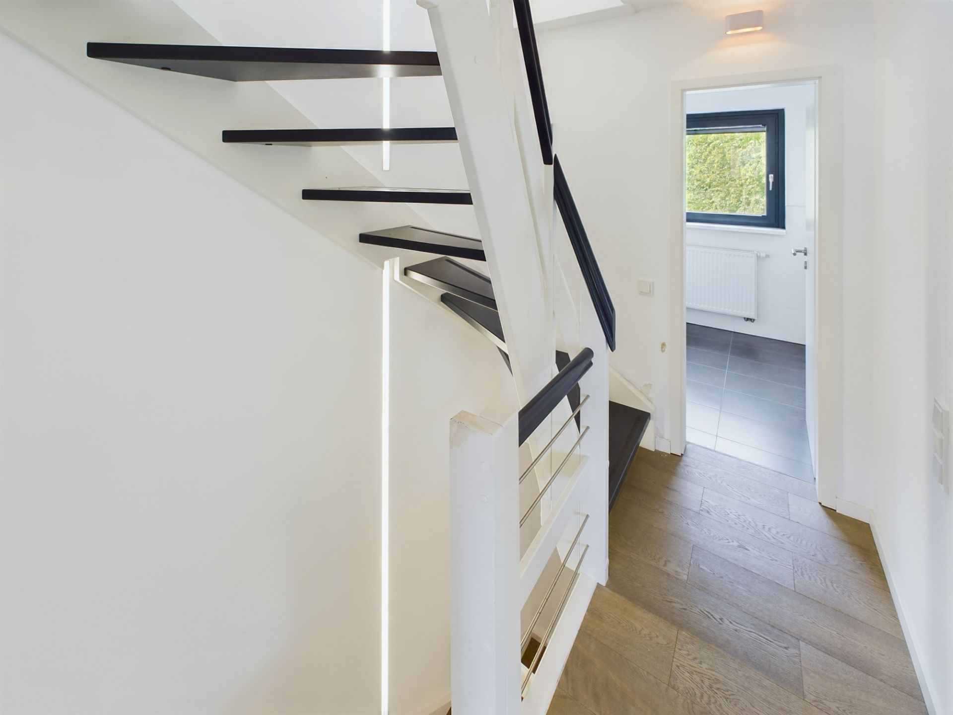Moderne Treppe mit schwarzem Geländer neben einem Flur mit Holzboden und Blick in ein Zimmer mit Fenster.