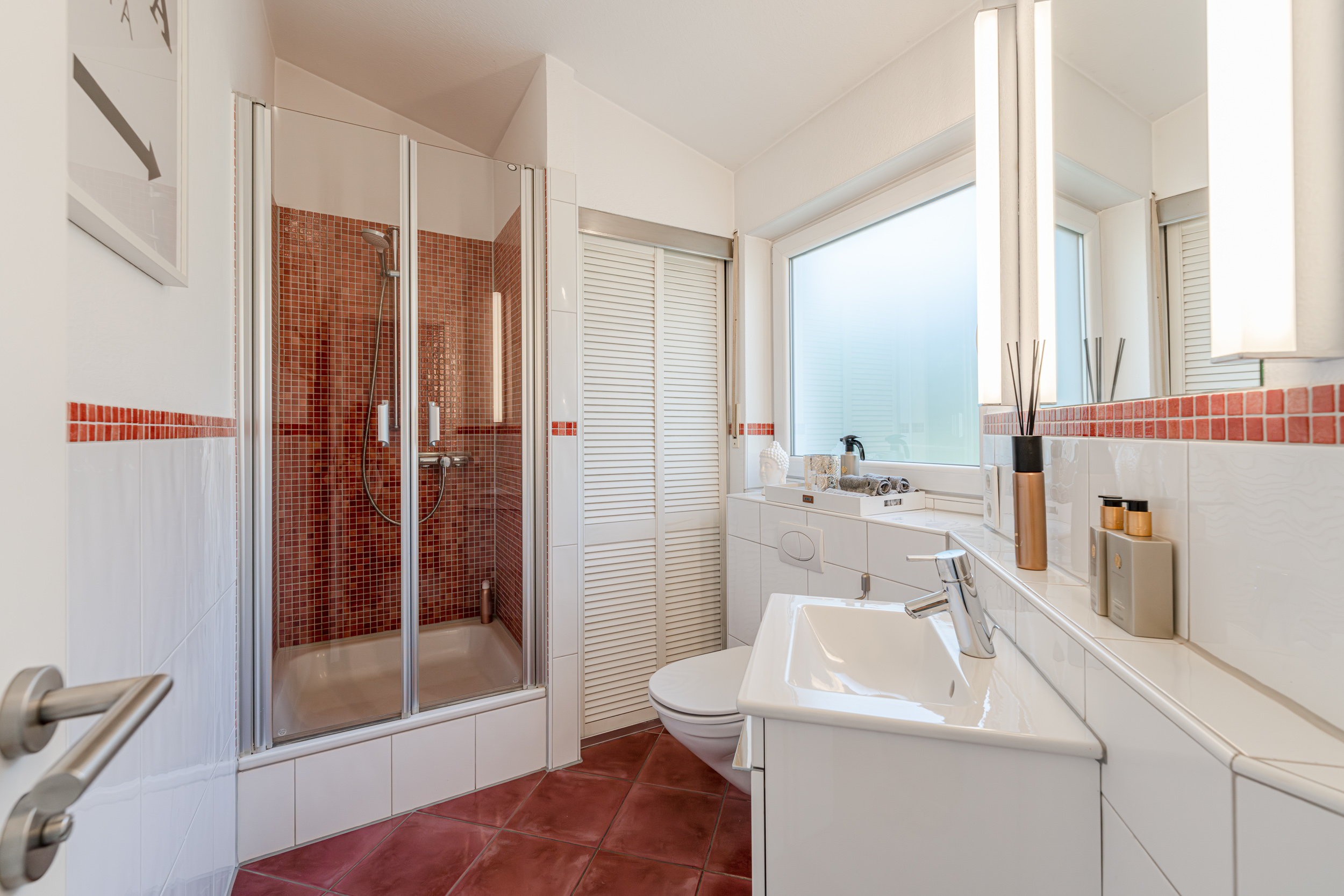 Modernes Badezimmer mit Dusche, Waschbecken und Toilette, rote Fliesen und großes Fenster.