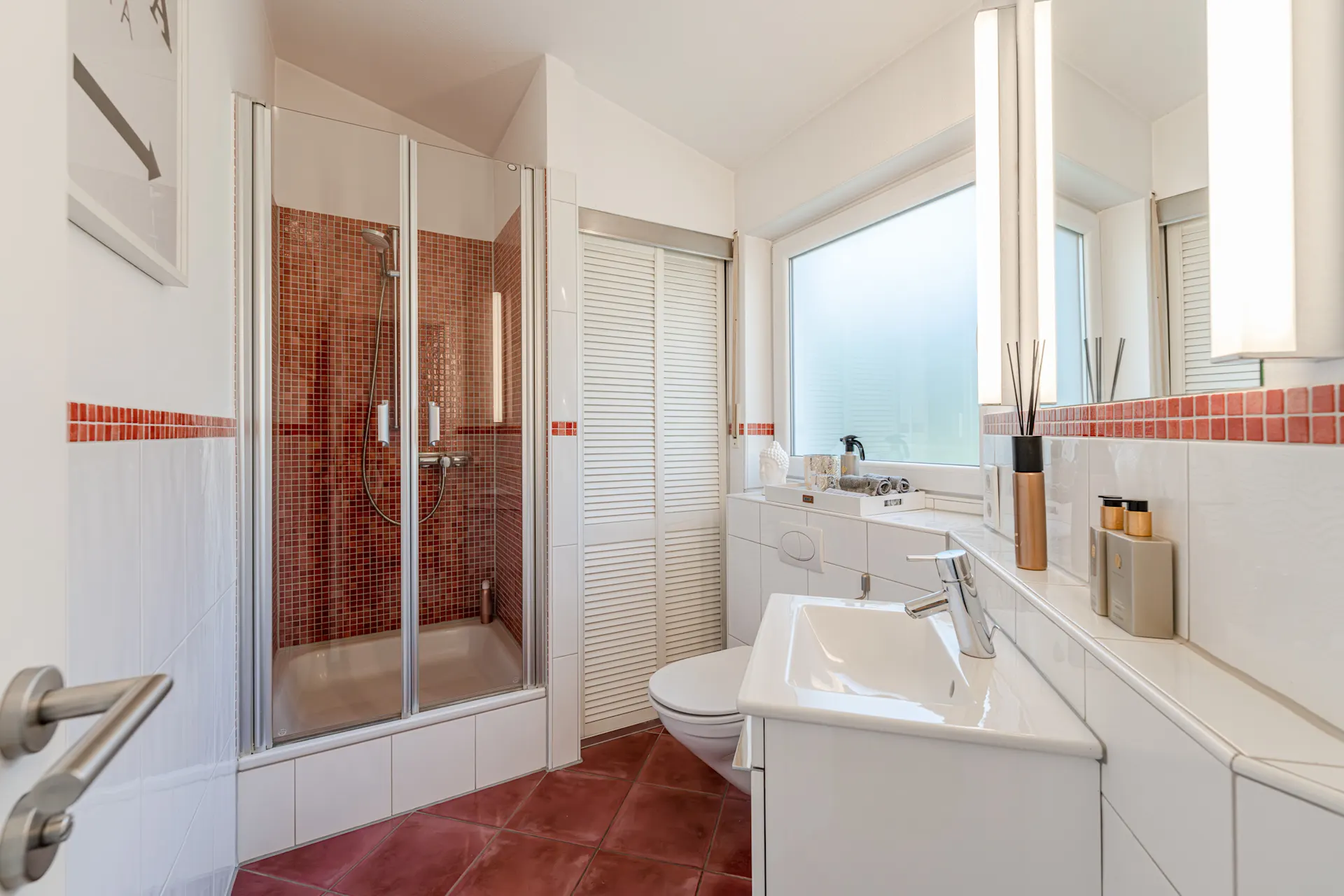 Modernes Badezimmer mit Dusche, Waschbecken und Toilette, rote Fliesen und großes Fenster.