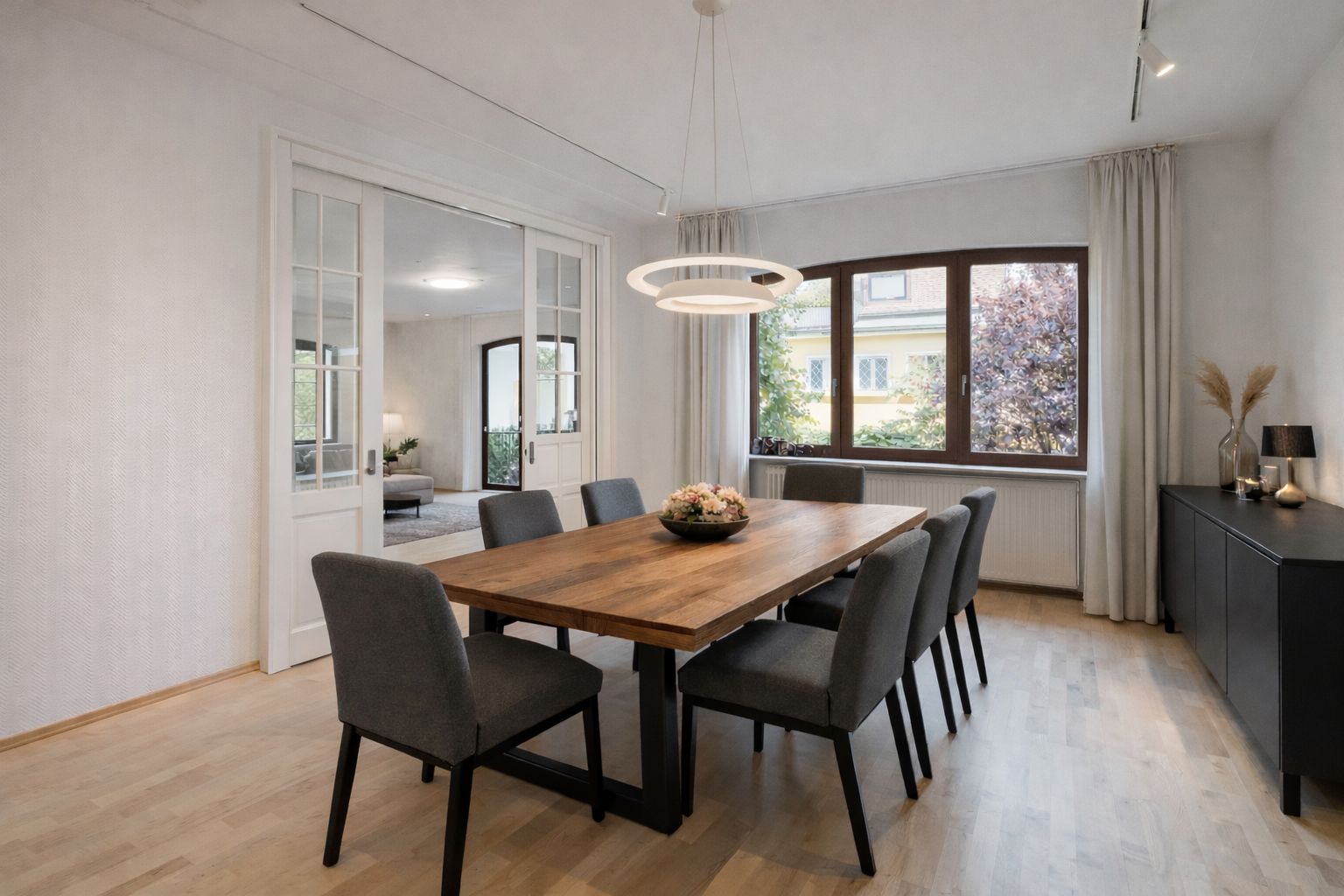 Esszimmer mit Holztisch, acht grauen Stühlen, großer Fensterfront und modernen Lampen.