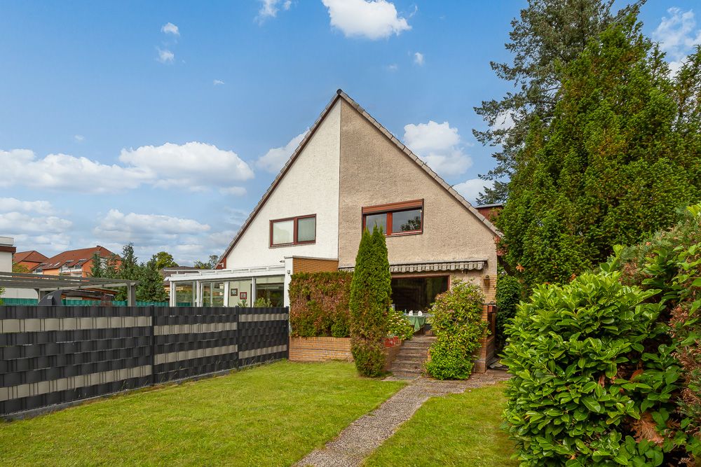 Zweistöckiges Haus mit spitzem Dach, Garten und Hecke, blauer Himmel im Hintergrund.