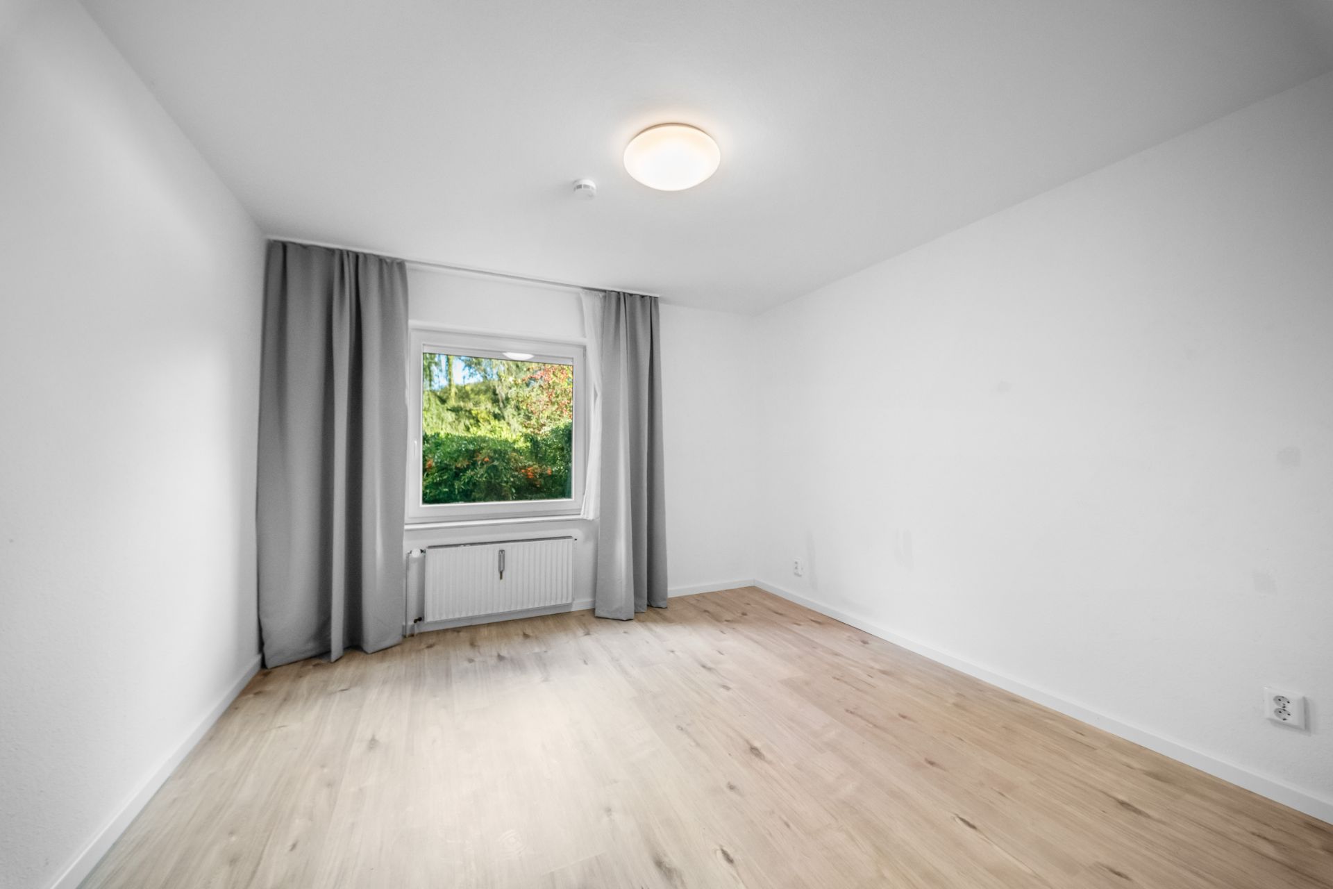 Helles, leeres Zimmer mit Holzboden, weißer Wand, Fenster mit grauen Vorhängen und Deckenlampe.