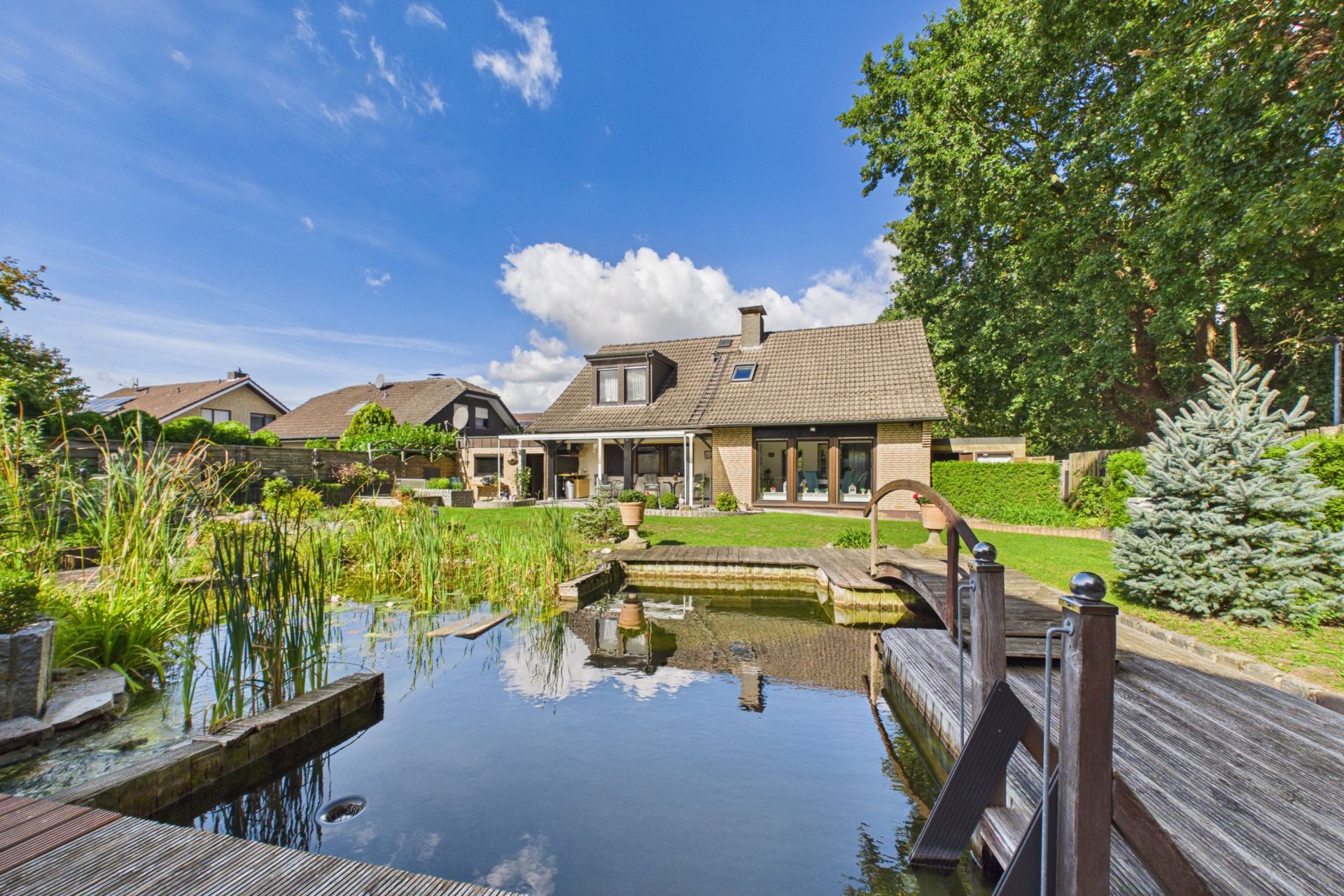 Großes Einfamilienhaus mit Garten, Teich und Holzsteg, umgeben von Bäumen unter blauem Himmel.