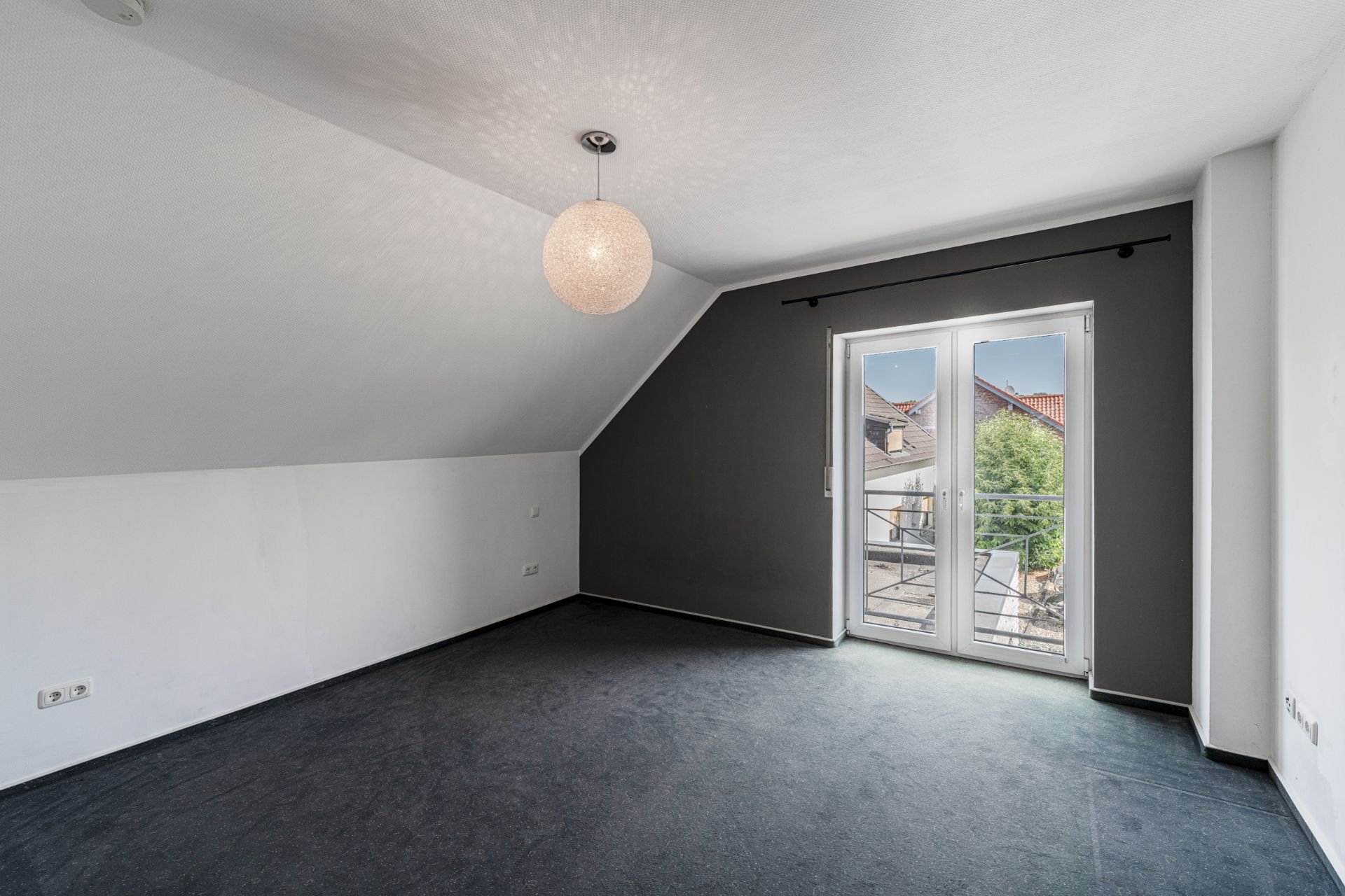 Leeres Zimmer mit Teppichboden, schräger Decke, runder Lampe und Doppeltür zum Balkon.