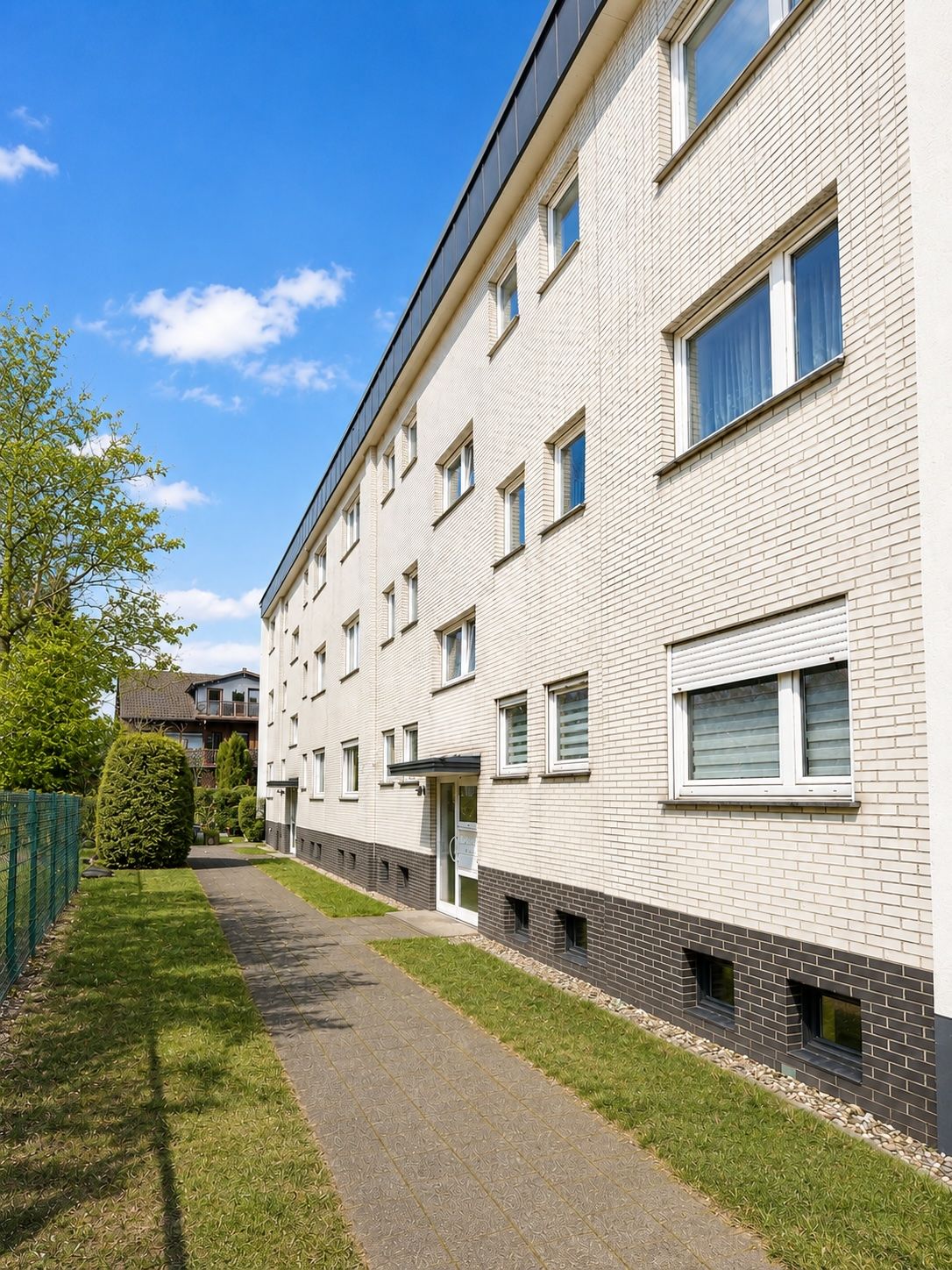 Mehrfamilienhaus mit hellem Klinker, mehreren Fenstern und Eingangsbereich, umgeben von Grünfläche.