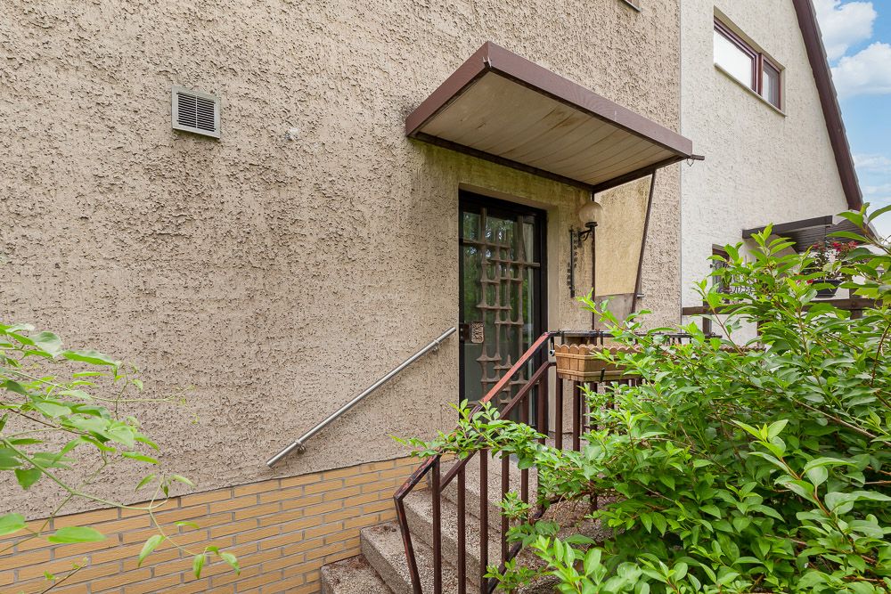 Eingangstür eines Wohnhauses mit Treppe, Geländer und Überdachung, umgeben von grünen Sträuchern.