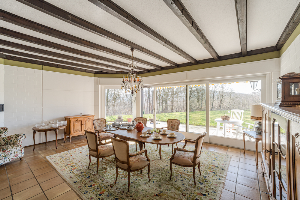 Heller Essbereich mit Holzbalkendecke, rundem Tisch, Teppich und großen Fenstern zum Garten.