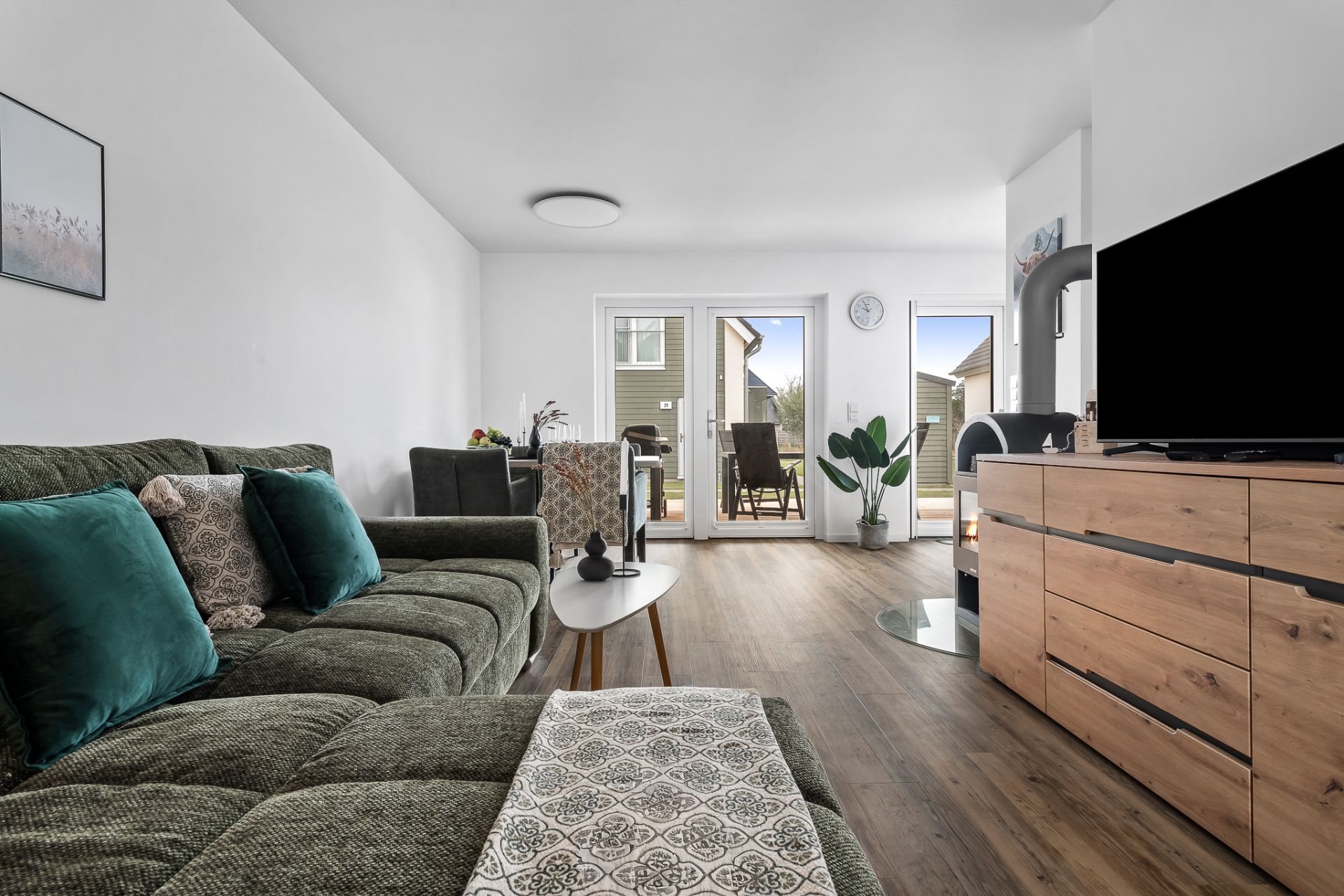 Modernes Wohnzimmer mit grünem Sofa, Holzkommode, Fernseher und Zugang zur Terrasse.