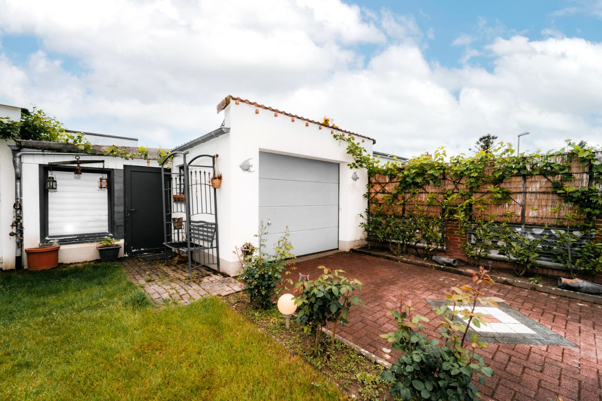 Garage mit weißem Tor, gepflasterter Einfahrt, Gartenzaun und angrenzendem Garten mit Rasen und Pflanzen.