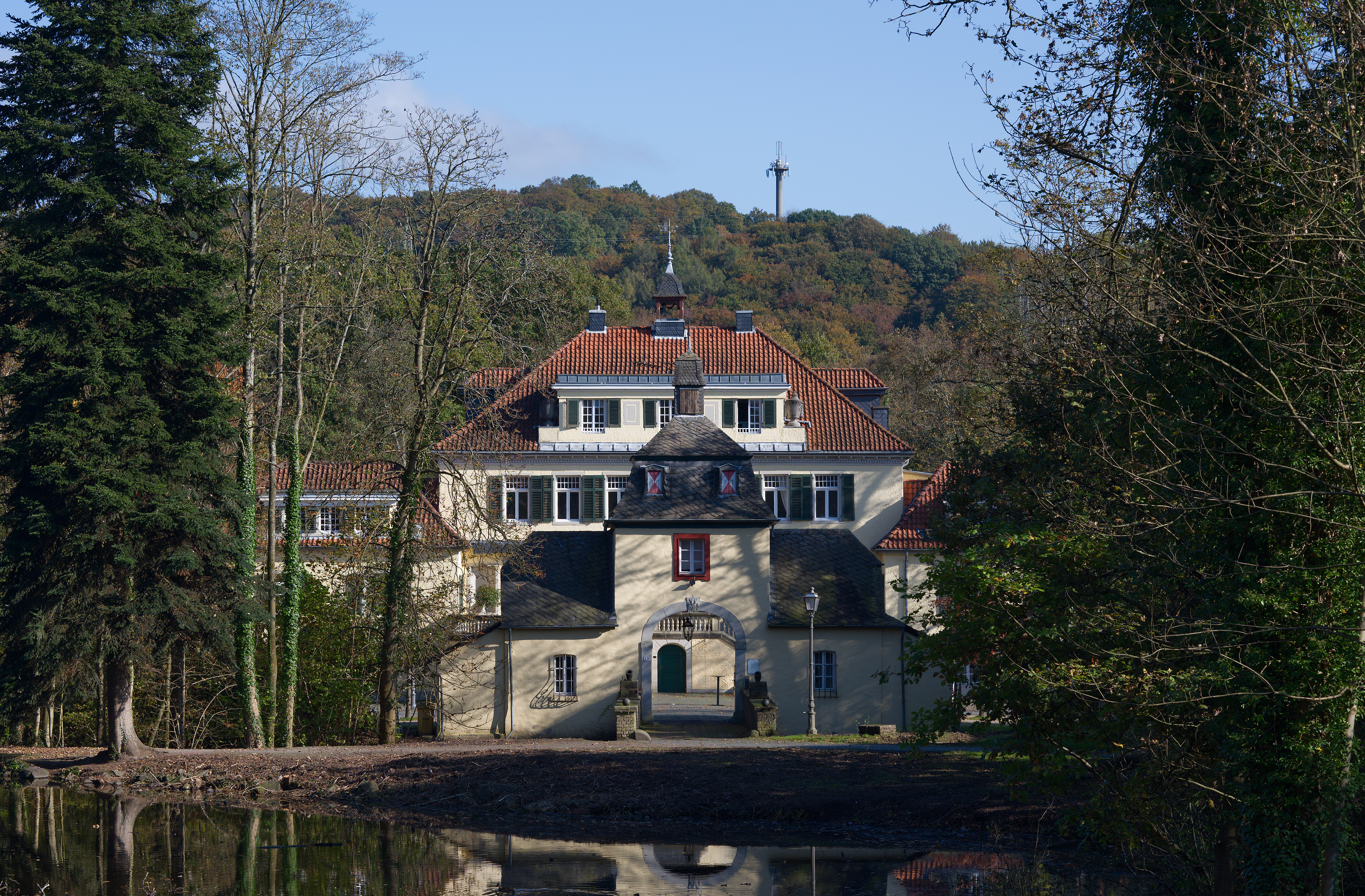 Großes Wohngebäude mit rotem Dach, umgeben von Bäumen und einem Teich im Vordergrund.
