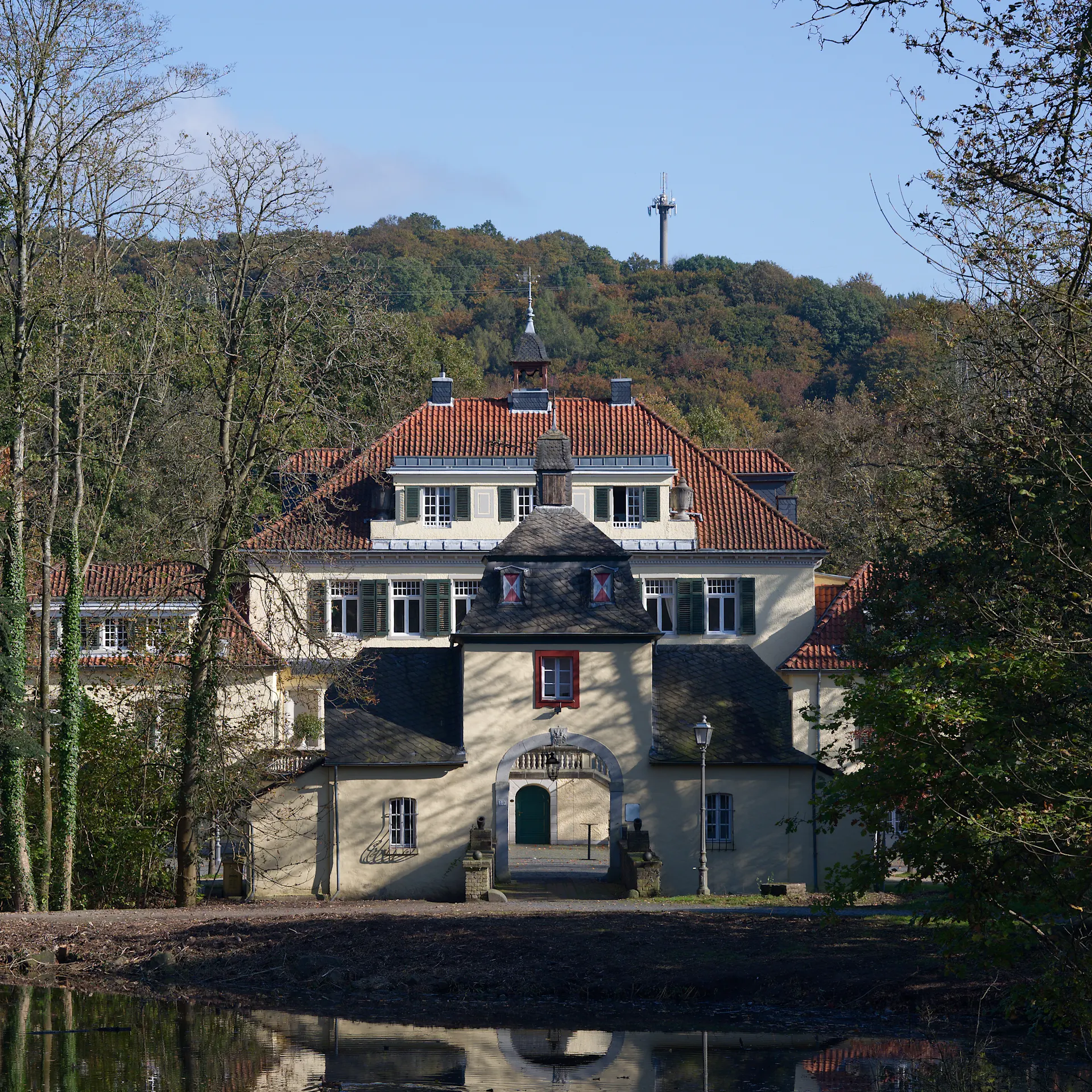 Großes Wohngebäude mit rotem Dach, umgeben von Bäumen und einem Teich im Vordergrund.