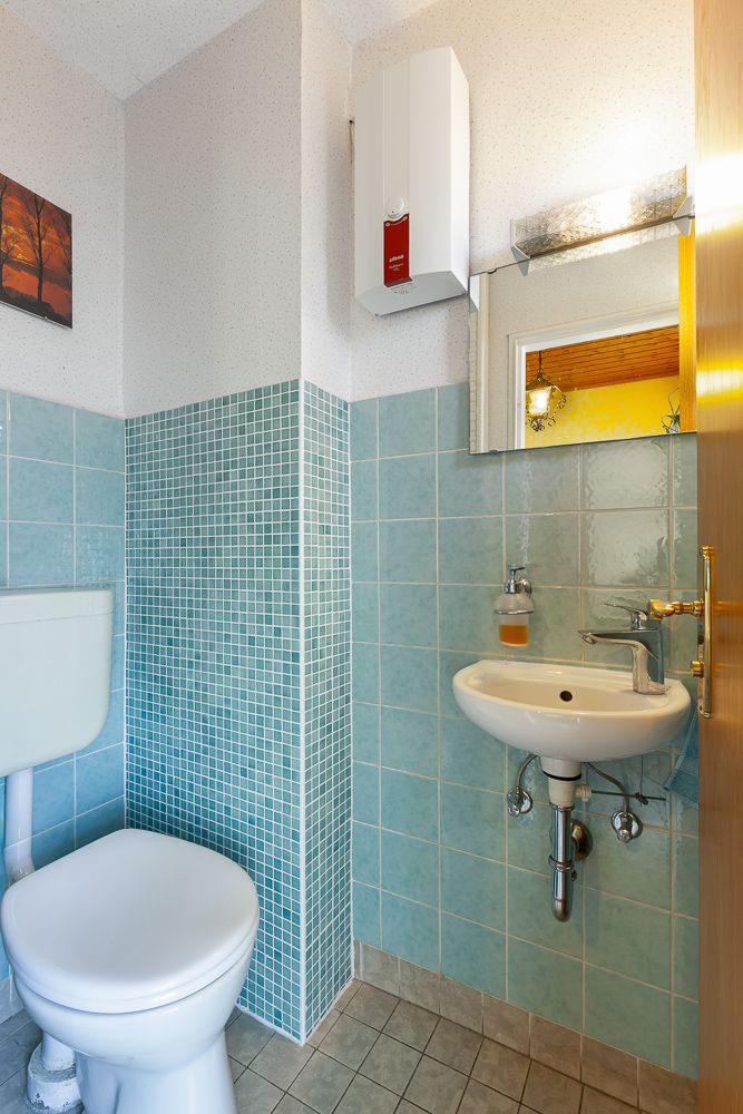 Kleines Badezimmer mit Toilette, Waschbecken und Wandspiegel, blaue Fliesen an den Wänden.