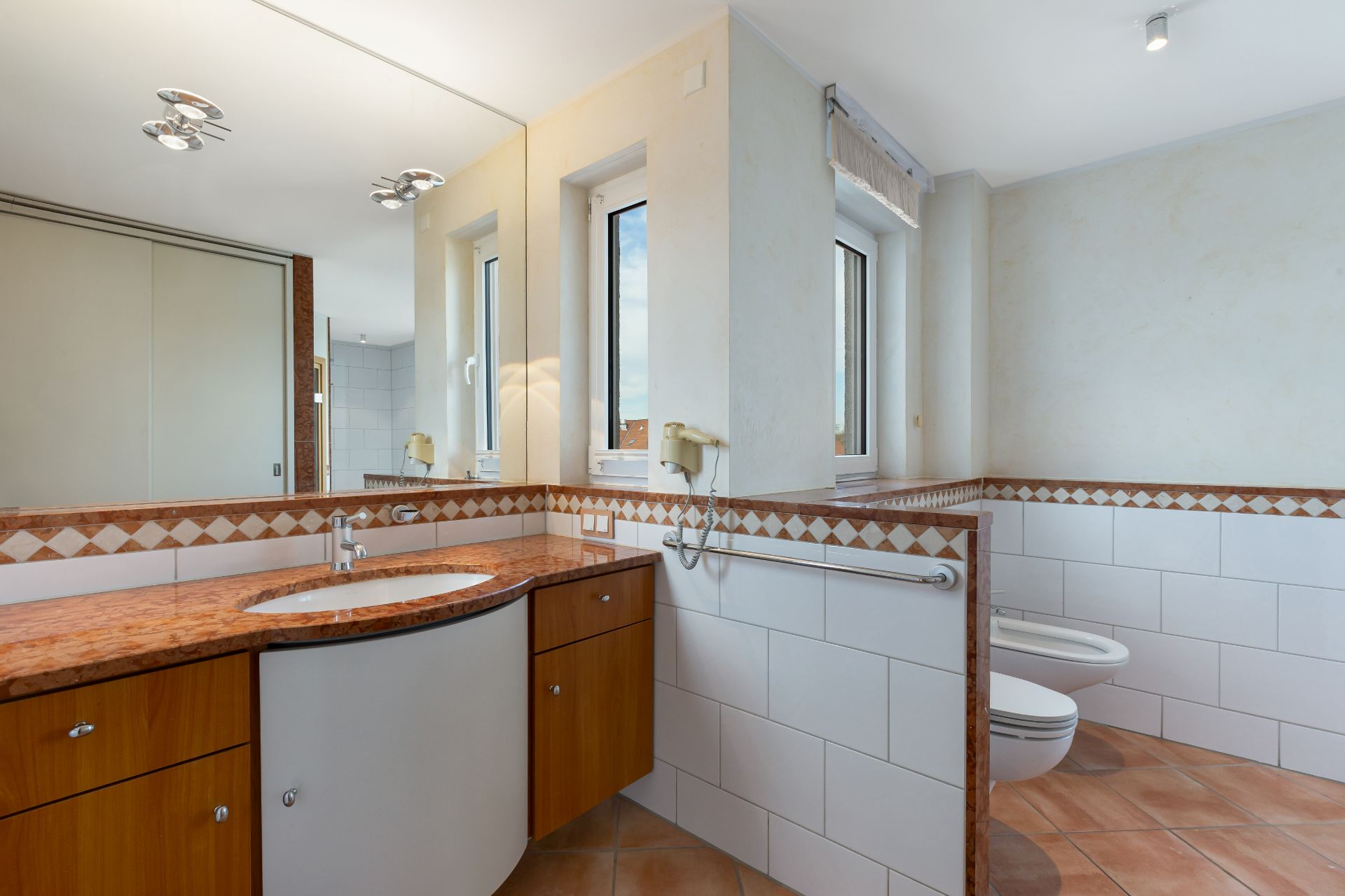 Modernes Badezimmer mit großem Spiegel, Waschbecken, Holzschränken und zwei Fenstern.