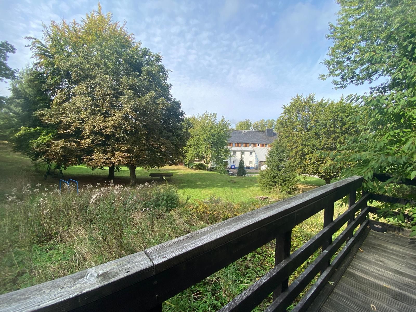 Blick von einer Holzterrasse auf einen Garten mit Bäumen und einem Wohnhaus im Hintergrund.