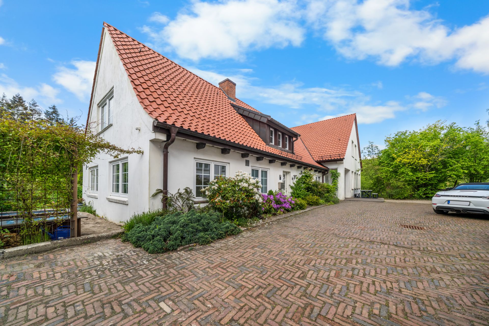 Weißes Wohnhaus mit rotem Ziegeldach, großem Hof und Blumenbeeten vor dem Eingang.