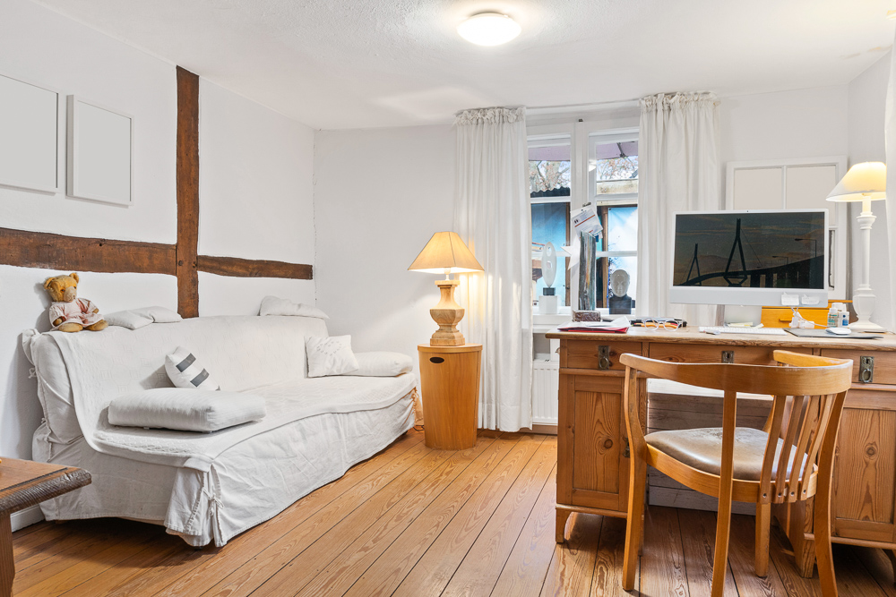 Wohnzimmer mit weißem Sofa, Holzboden, Schreibtisch, Computer und großen Fenstern.