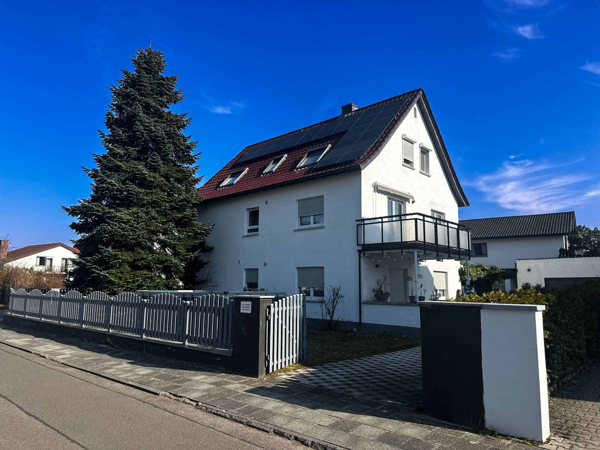 Weißes Wohnhaus mit rotem Dach, Balkon, Solarpanels und großem Baum im Vorgarten.