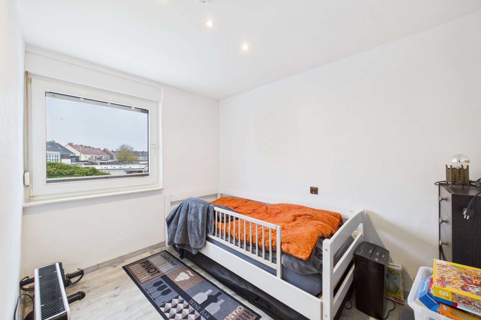 Kleines Schlafzimmer mit Einzelbett, Fenster, Teppich und wenigen Möbeln.
