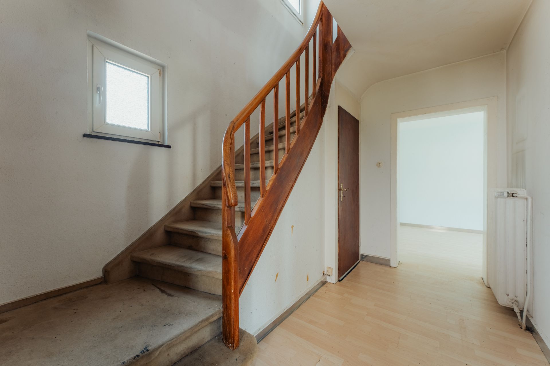 Flur mit Holztreppe, kleinem Fenster und offener Tür zu einem hellen Zimmer.