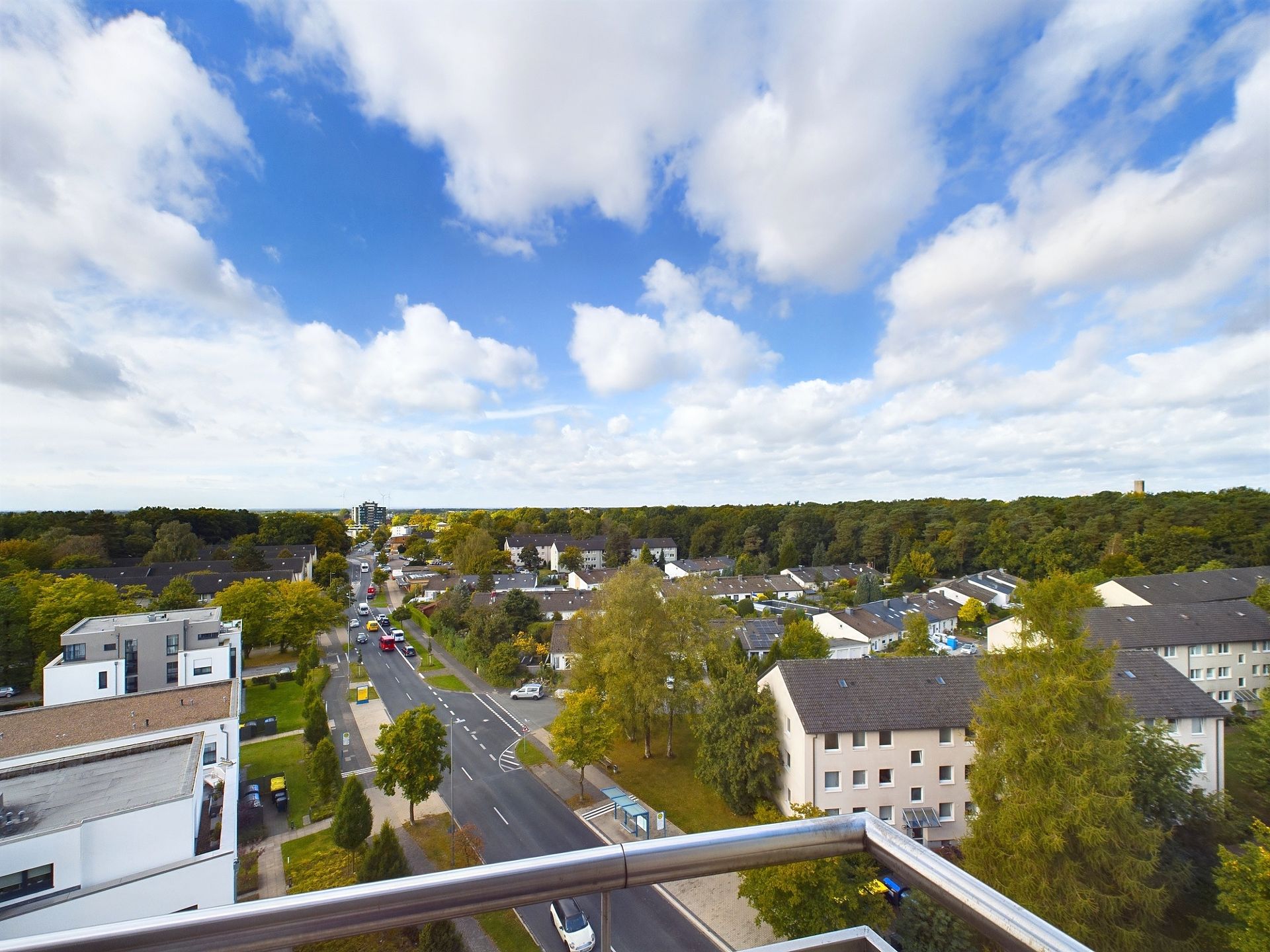 Aussicht von einem Balkon auf Wohnhäuser, eine Straße und viele Bäume bei blauem Himmel mit Wolken.