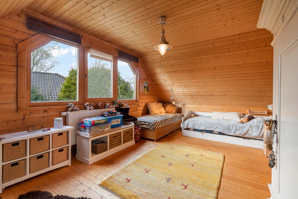 Helles Kinderzimmer mit Holzdecke, Spielzeugregal, zwei Betten und großem Fenster.